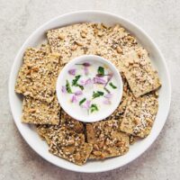 A plate of multi-seed oat flour crackers on a small white plate with a bowl of lemon herb yogurt dip in the middle of the plate.
