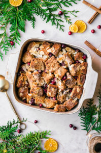 White wine bread pudding in a baking dish surrounded by greenery, oranges, and cinnamon sticks.