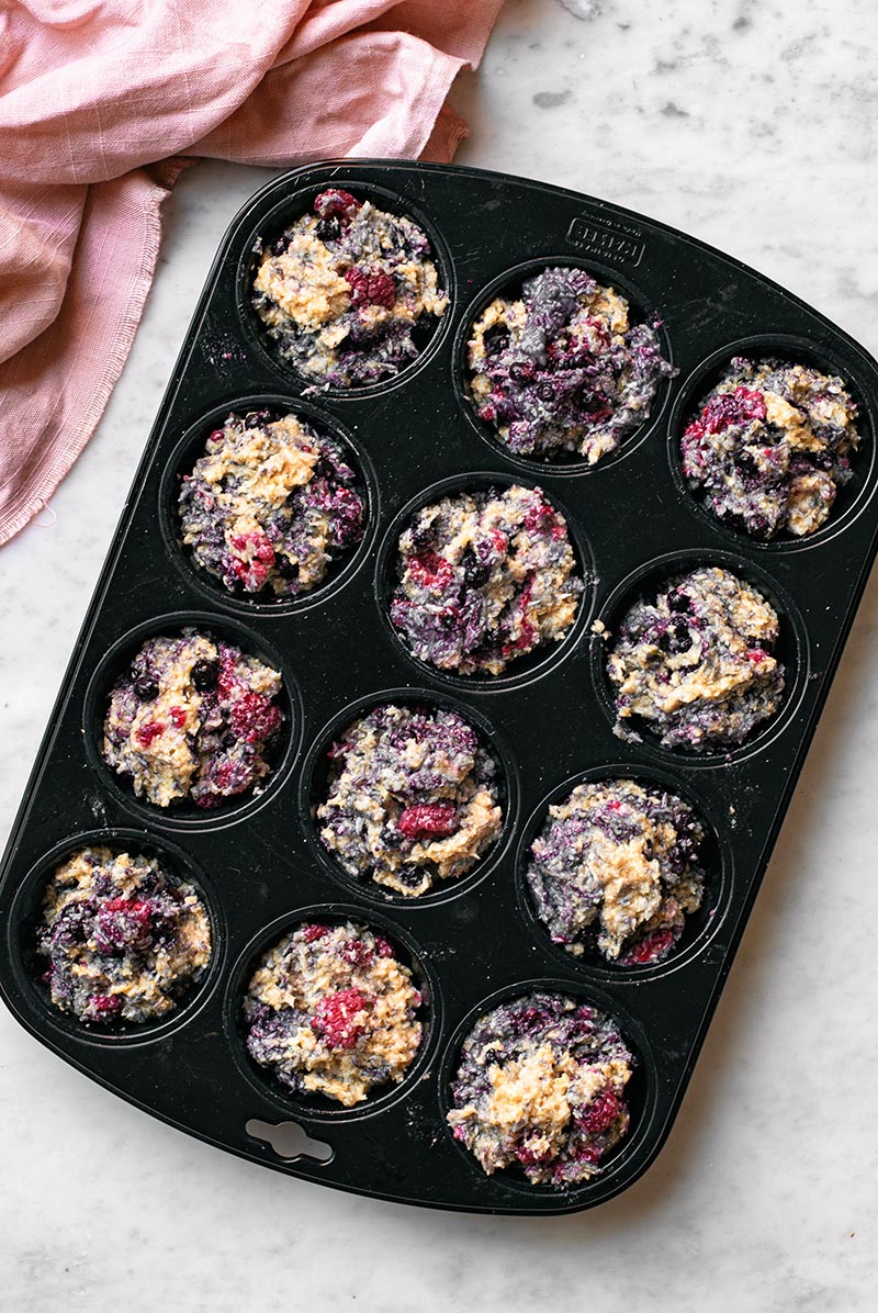 A muffin pan filled with batter for mixed berry muffins with toasted coconut.