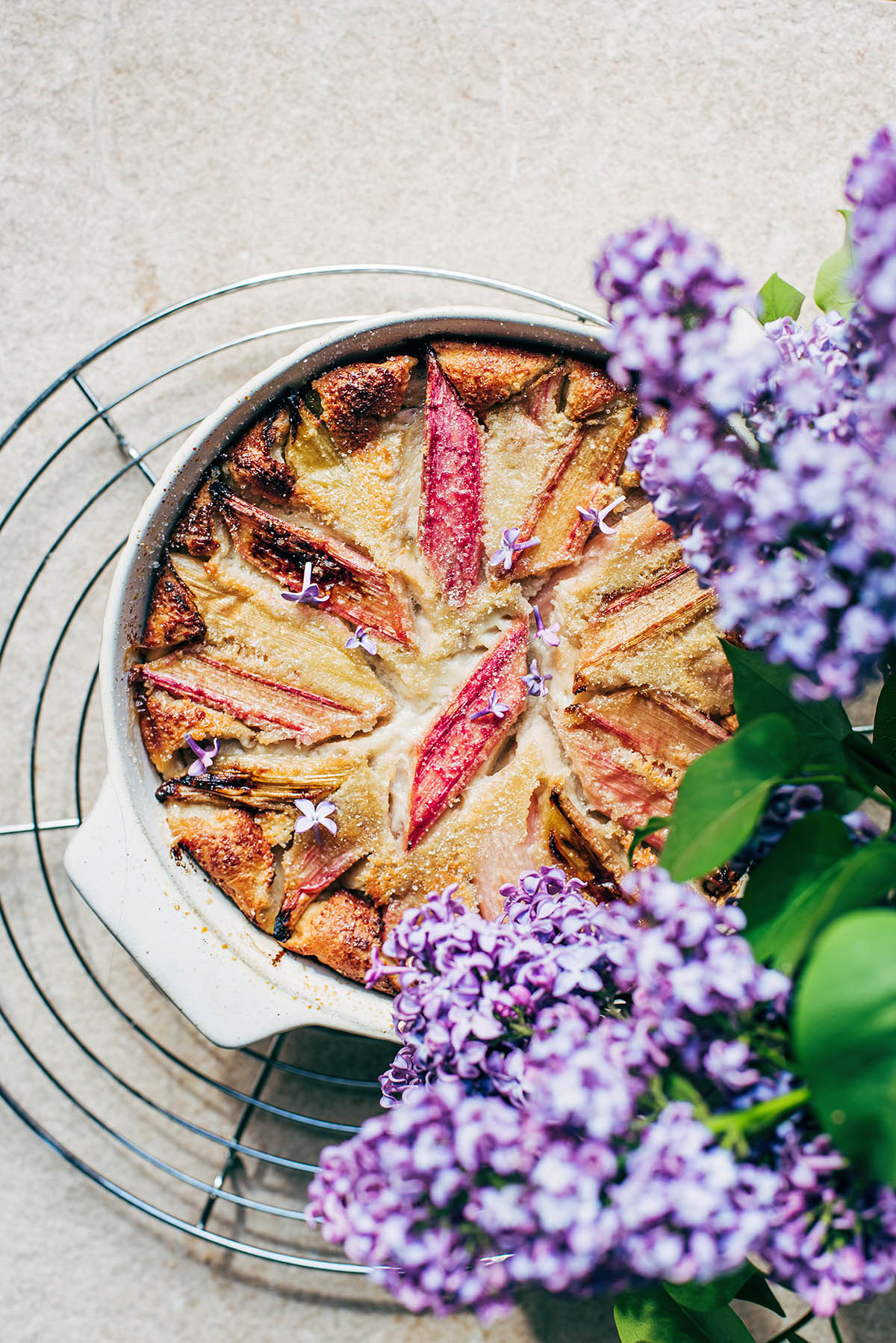 A rhubarb clafoutis on a cooling rack with lilac flowers in the foreground.