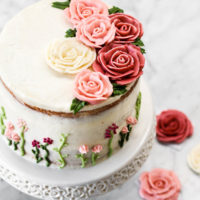 A tall cake on a cake stand with buttercream roses on top and small flowers on the sides.