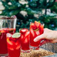 A hand taking a glass of strawberry lemonade sangria from a tray with two other filled glasses and a pitcher of sangria.
