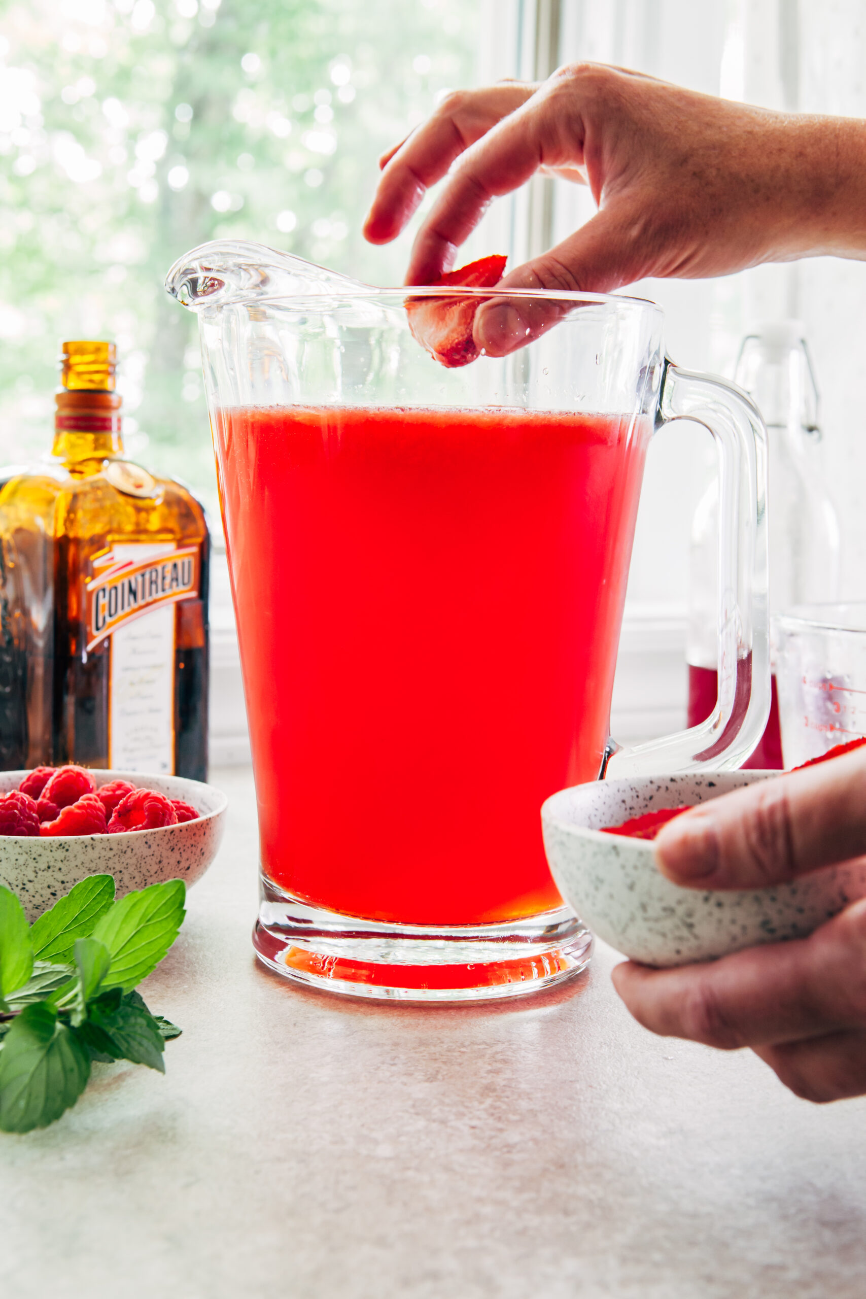 A woman's hand adding a sliced strawberry to a glass pitcher filled with sangria.