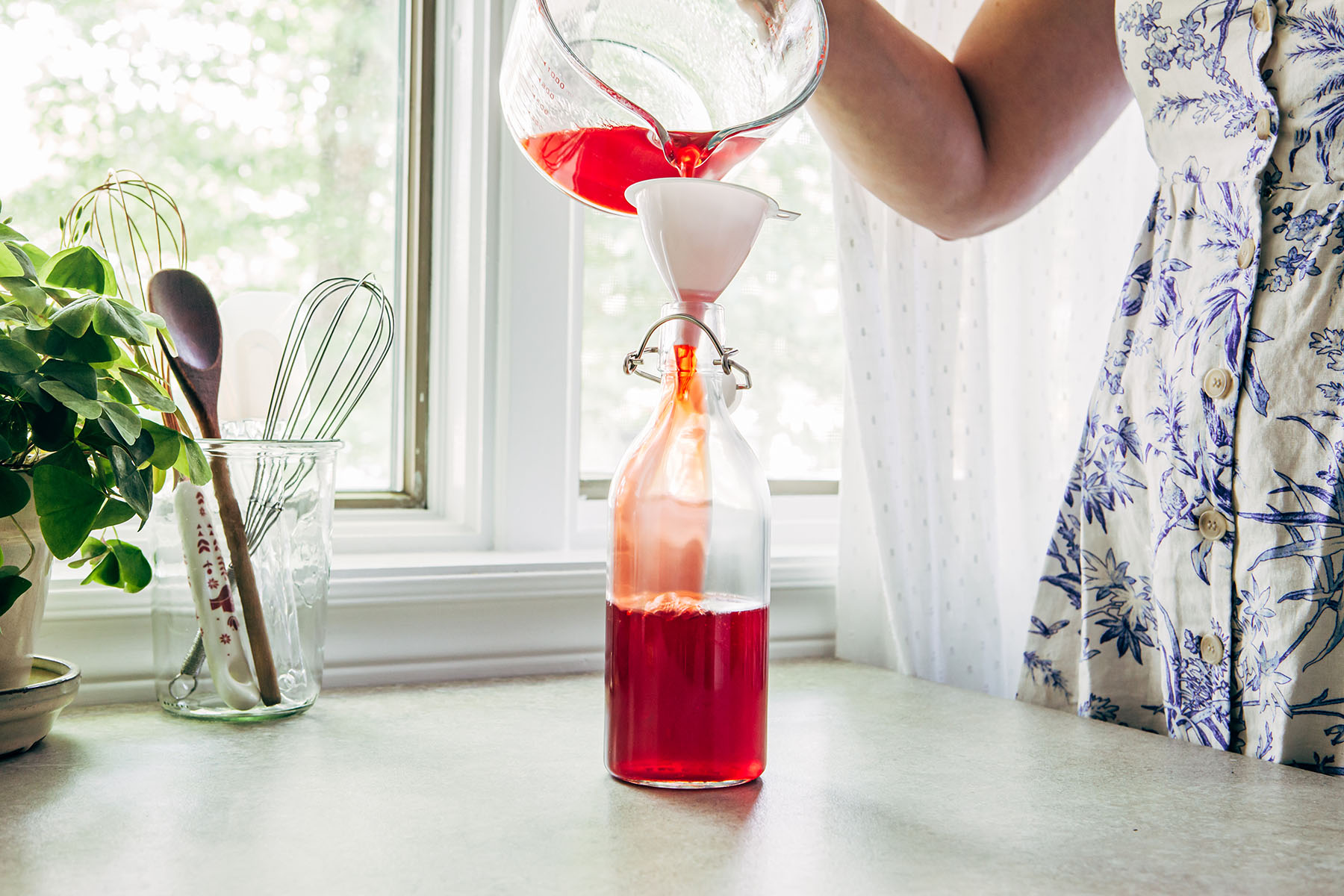 A woman in a blue and white dress funnelling strawberry ginger simple syrup into a glass bottle with a flip top lid.