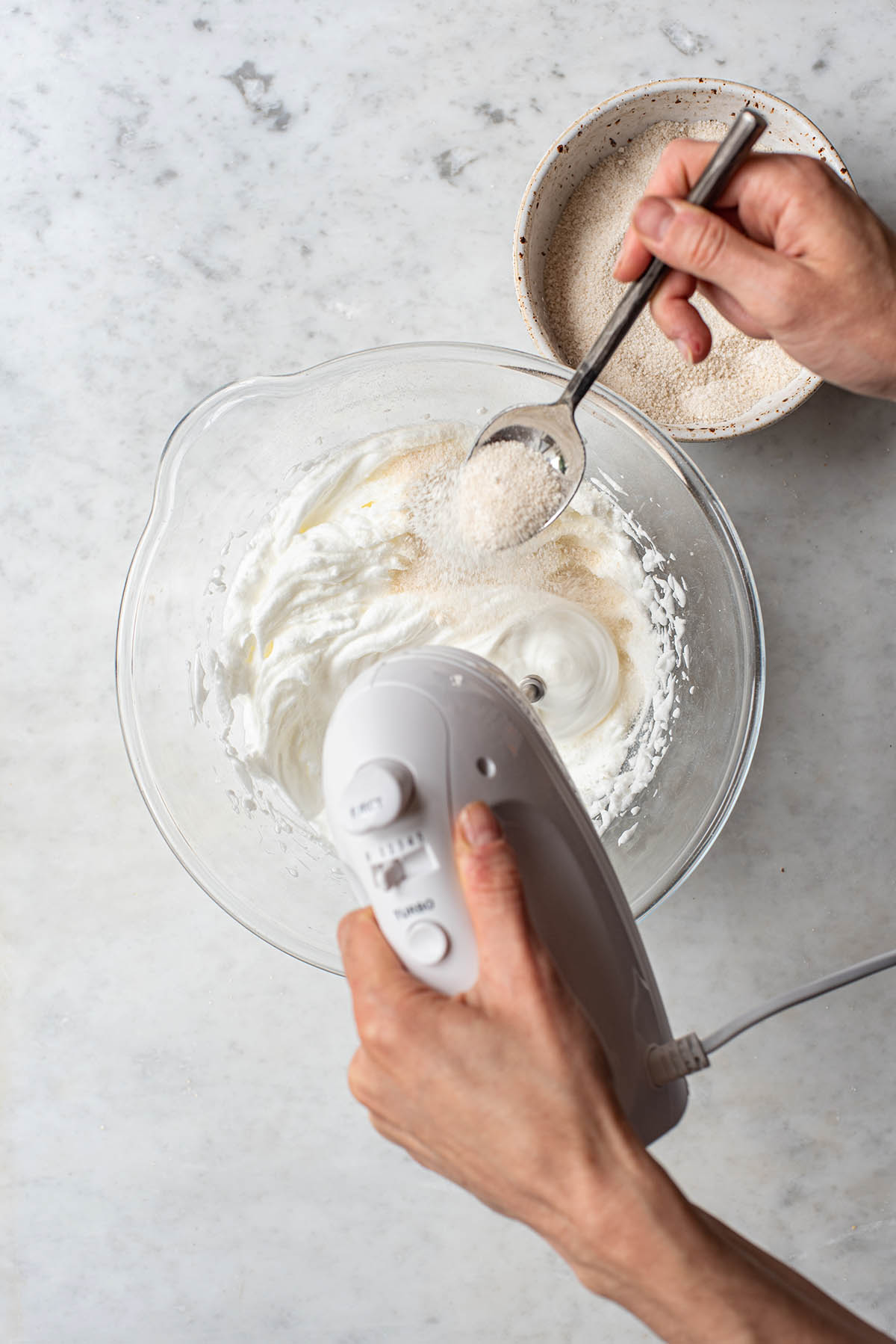 A hand adding a spoonful of sugar to a bowl of meringue while using the other hand to mix with a hand mixer.