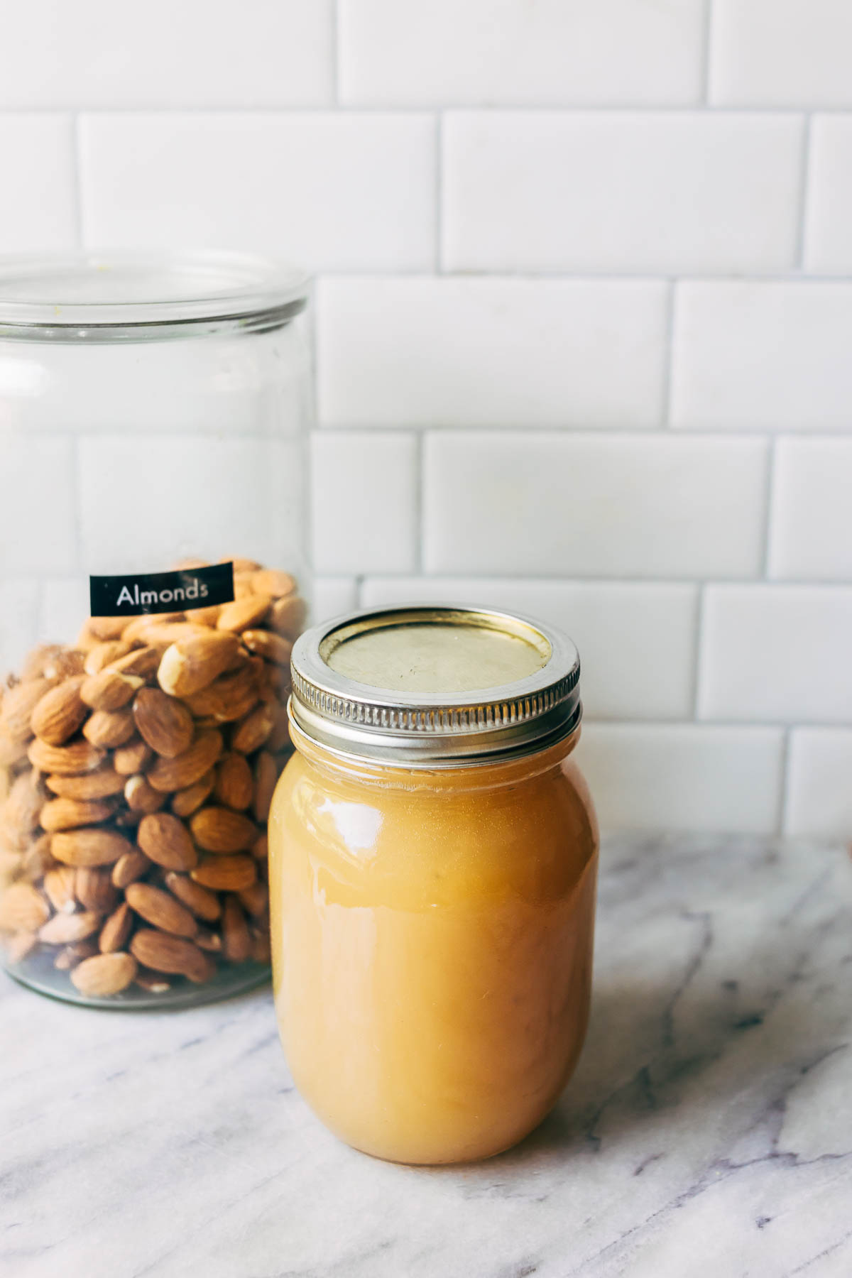 A jar of homemade orgeat syrup on a counter next to a glass canister half-filled with almonds.