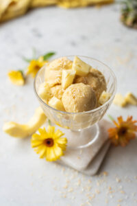A dish of coconut pineapple ice cream with chunks of fresh pineapple on top and yellow flowers scattered on the marble surface below.