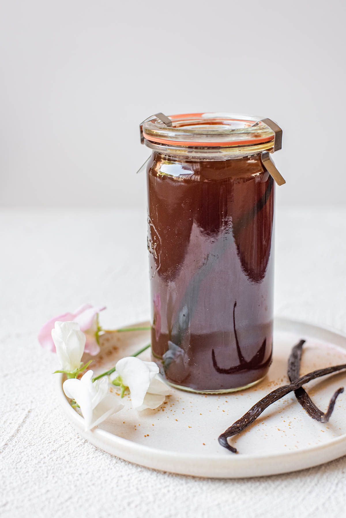 A jar of brown syrup with vanilla beans on a white plate on a white plaster surface with a few sweet pea flowers strewn about.