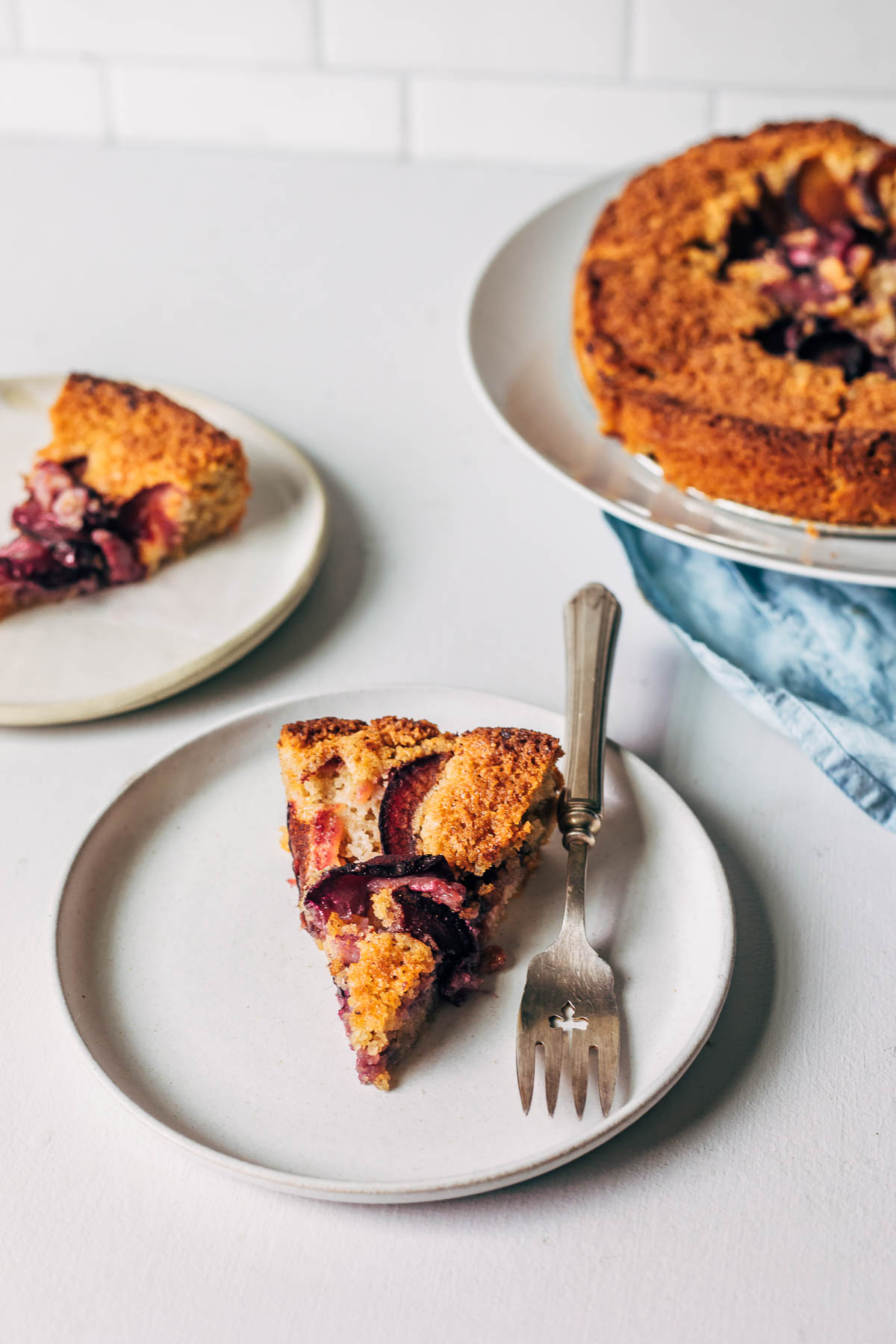 A slice of gluten-free plum cake on a plate with a fork next to another slice on a plate and the whole cake itself.