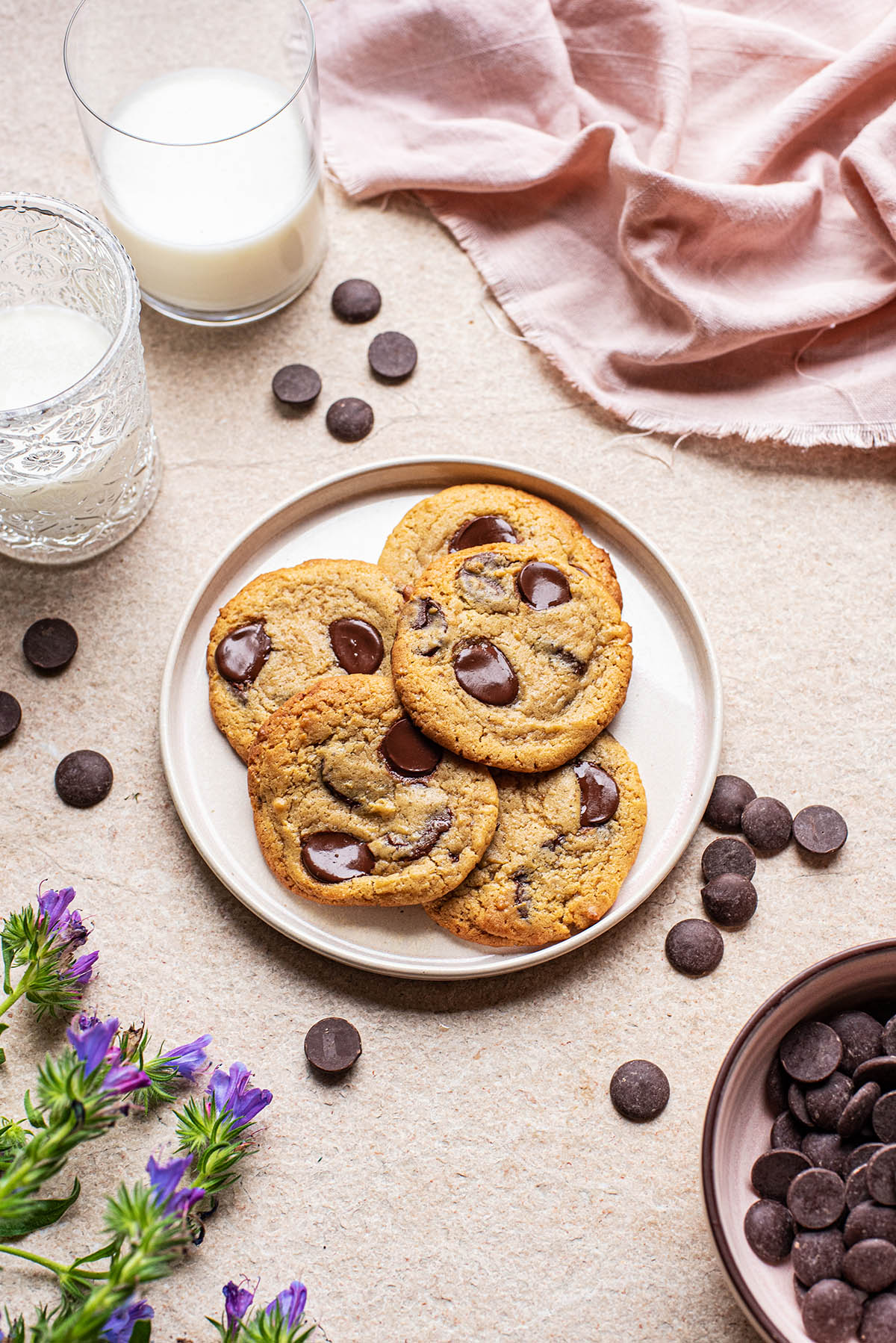A plate of chocolate chip cookies on a stone surface with a pink linen, glasses of milk, scattered chocolate chips, and purple flowers nearby.