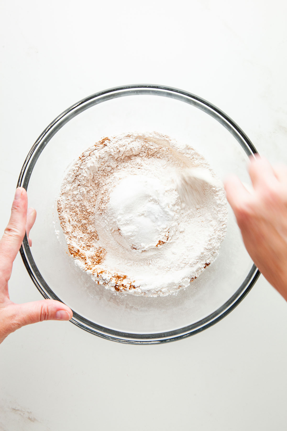 A hand mixing flour, sugar, and spice in a glass bowl.