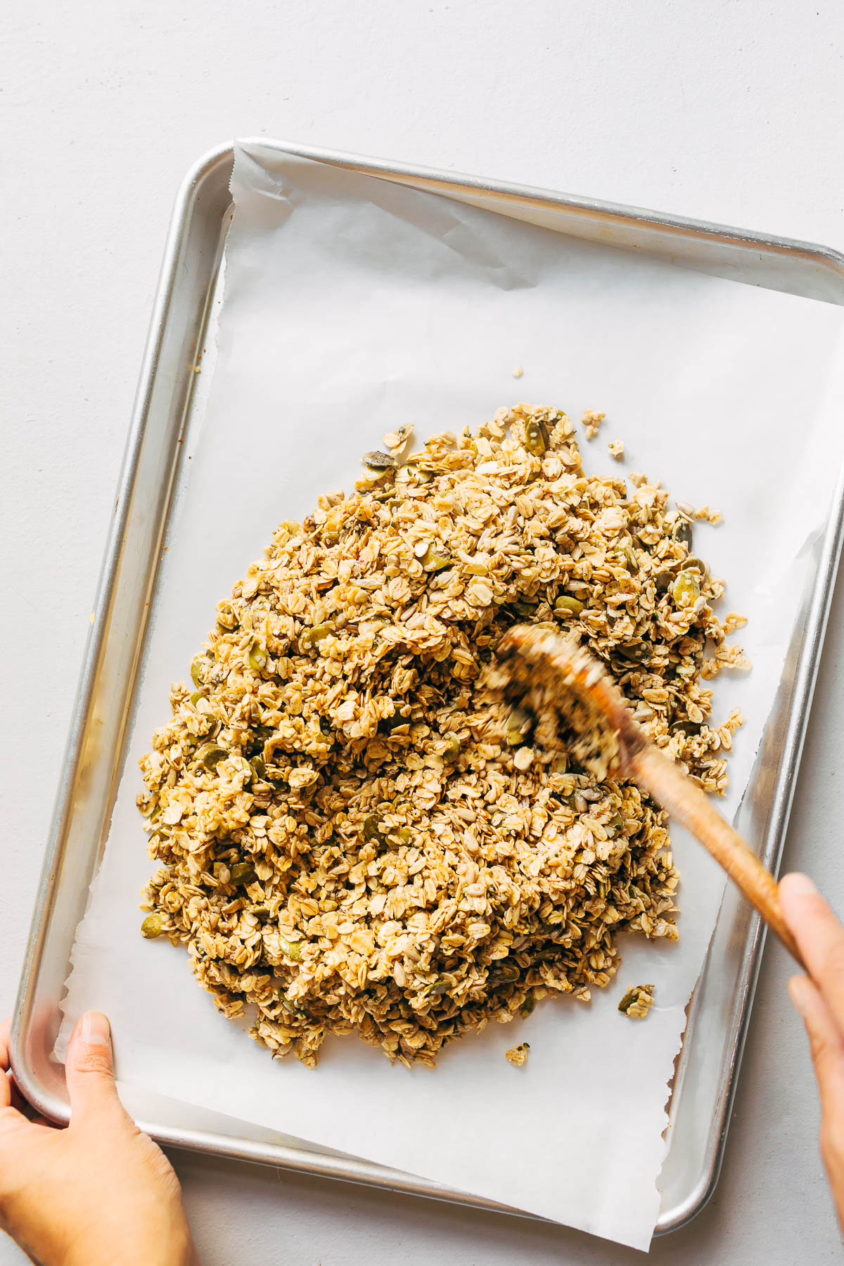 A hand spreading unbaked granola onto a parchment-paper lined baking sheet.