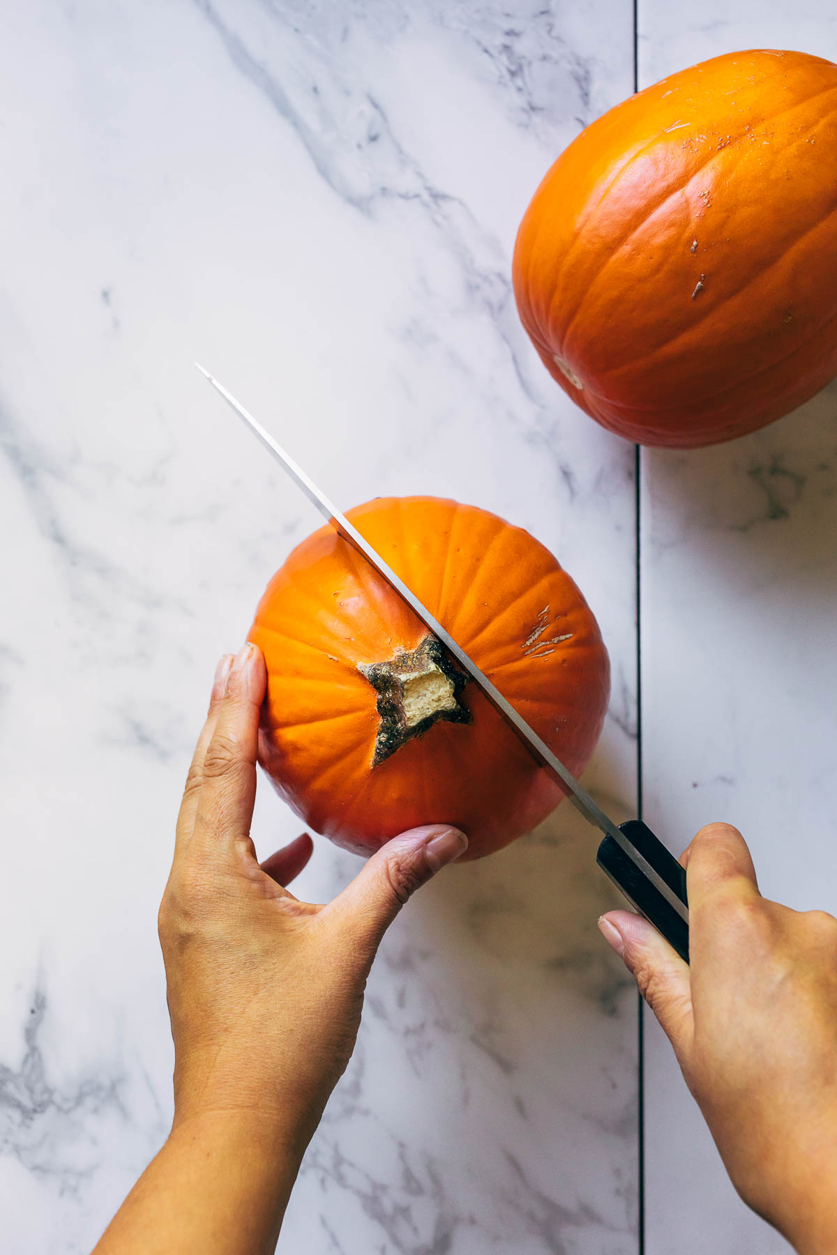 Hands slicing through a small pumpkin with a large chef's knife.