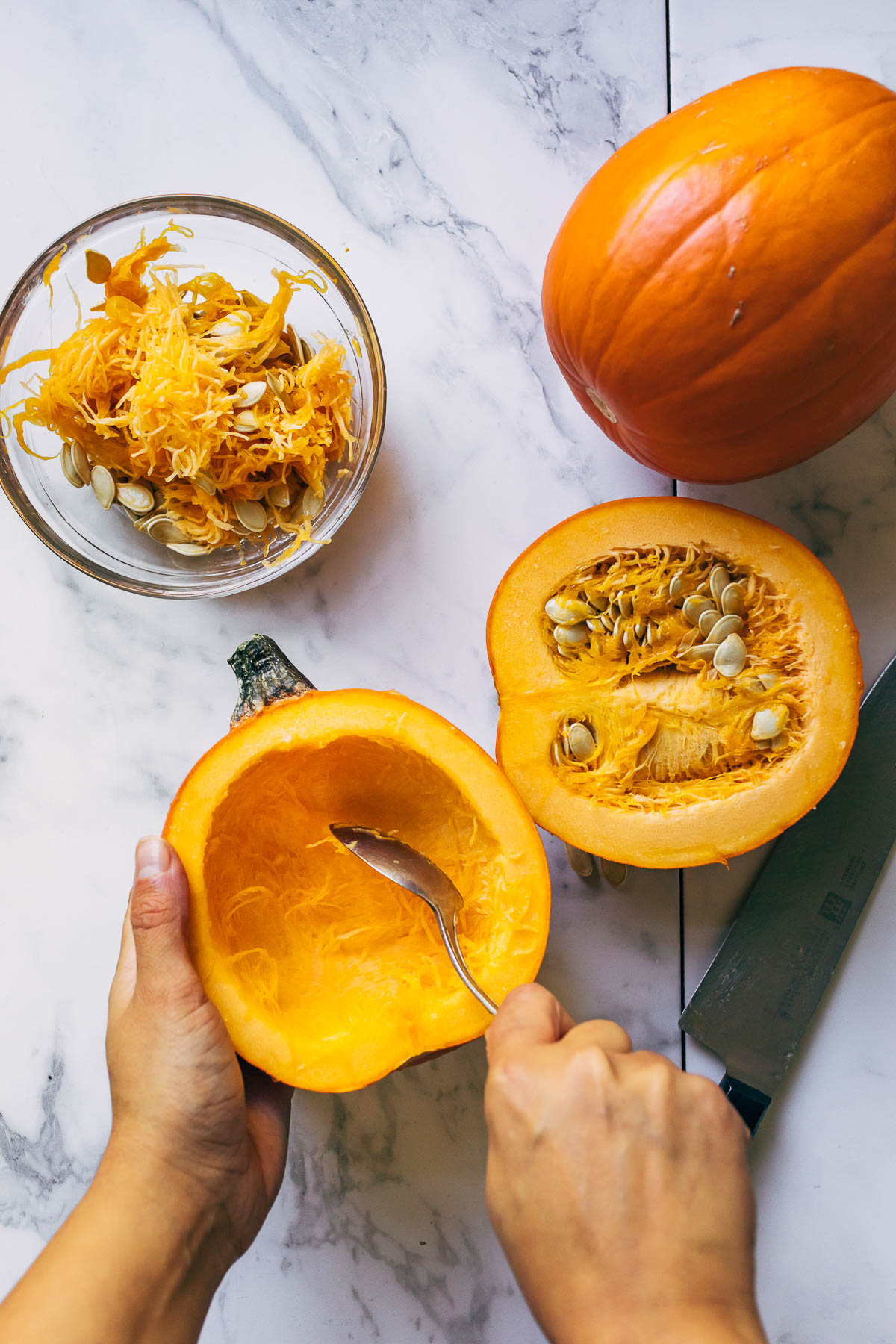 A hand scooping strings out of a small pumpkin with a spoon.