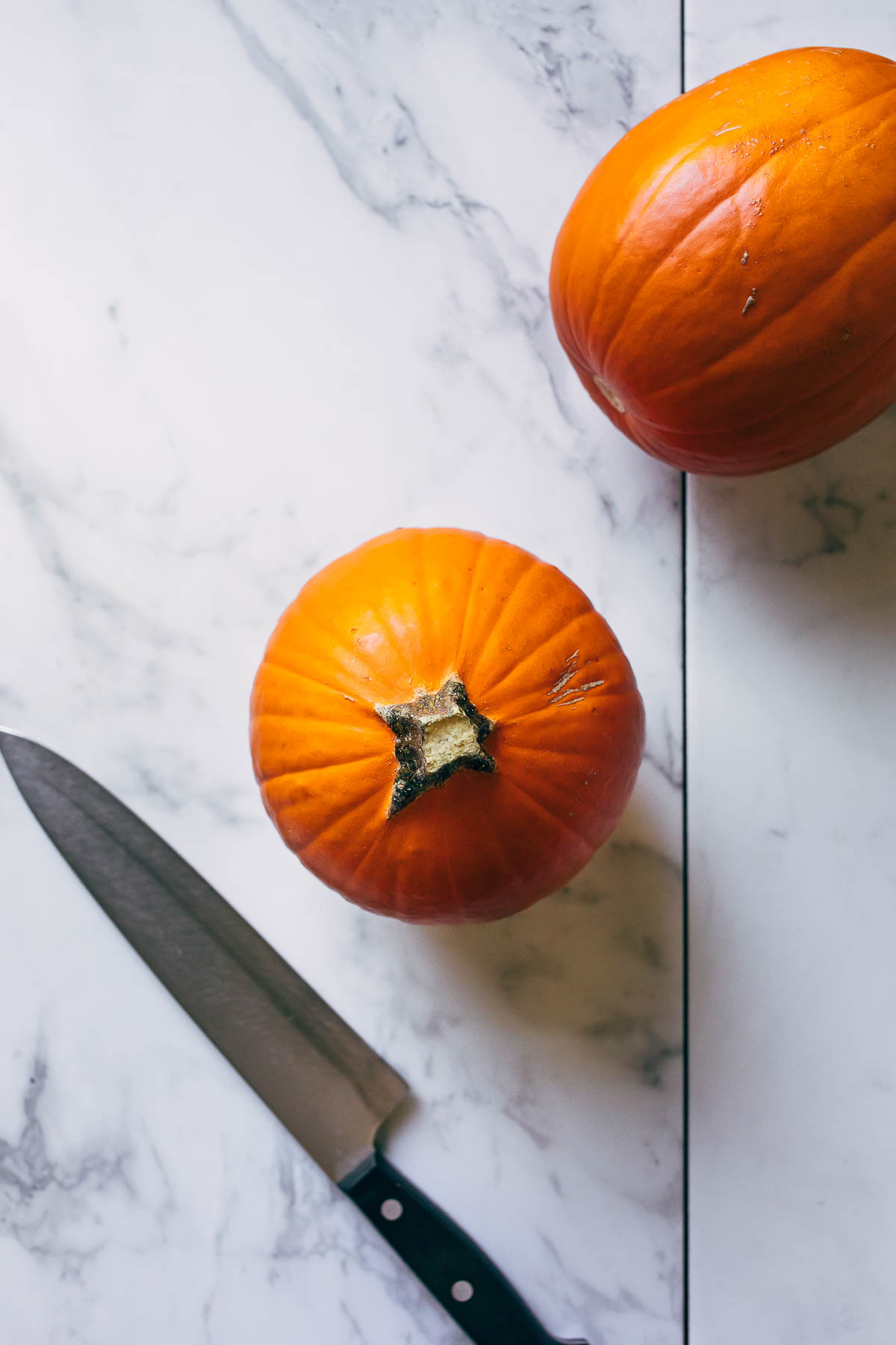 Two small sugar pumpkins on a marble surface alongside a chef's knife.