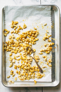 A metal baking tray of spicy roasted pumpkin seeds with parchment paper underneath and a spoon nearby.
