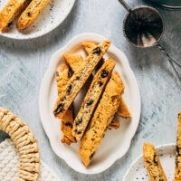 Chocolate chunk biscotti on a tray with more on plates and a cup of coffee.