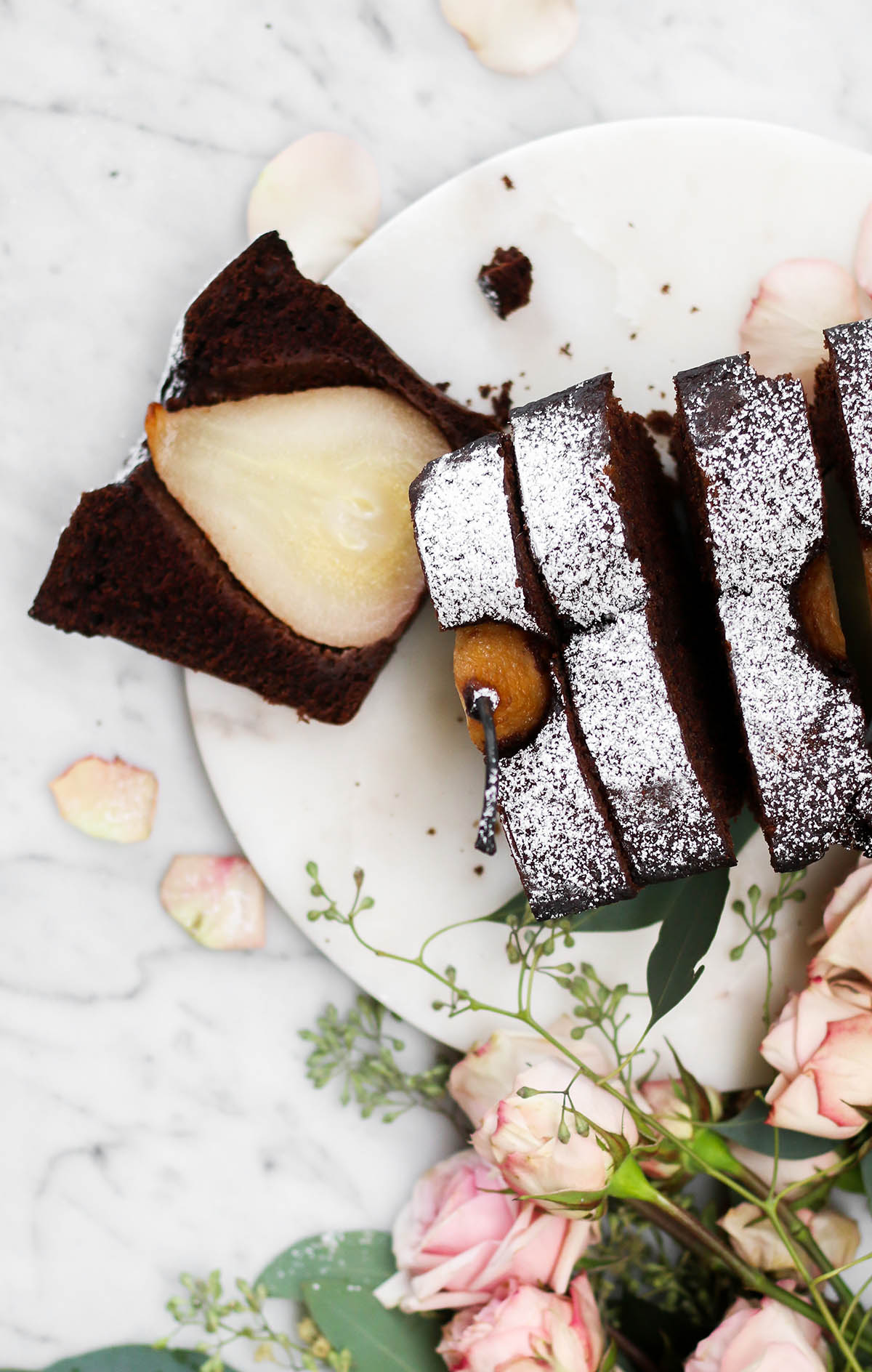A sliced chocolate loaf cake with pears on a white plate with light pink roses nearby.