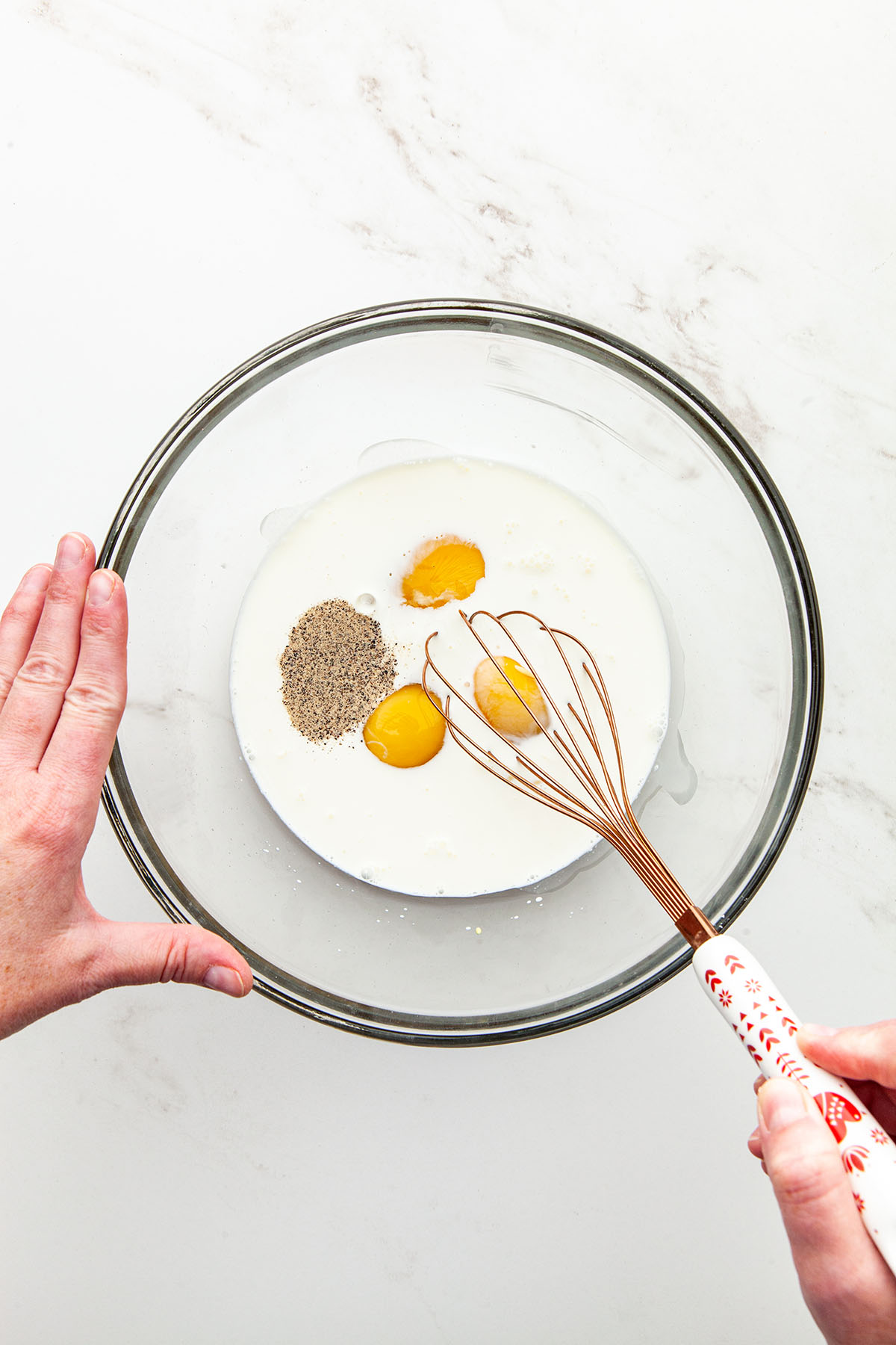 Hands using a whisk to mix eggs, cream, milk, salt, pepper, and Dijon mustard in a large glass bowl.