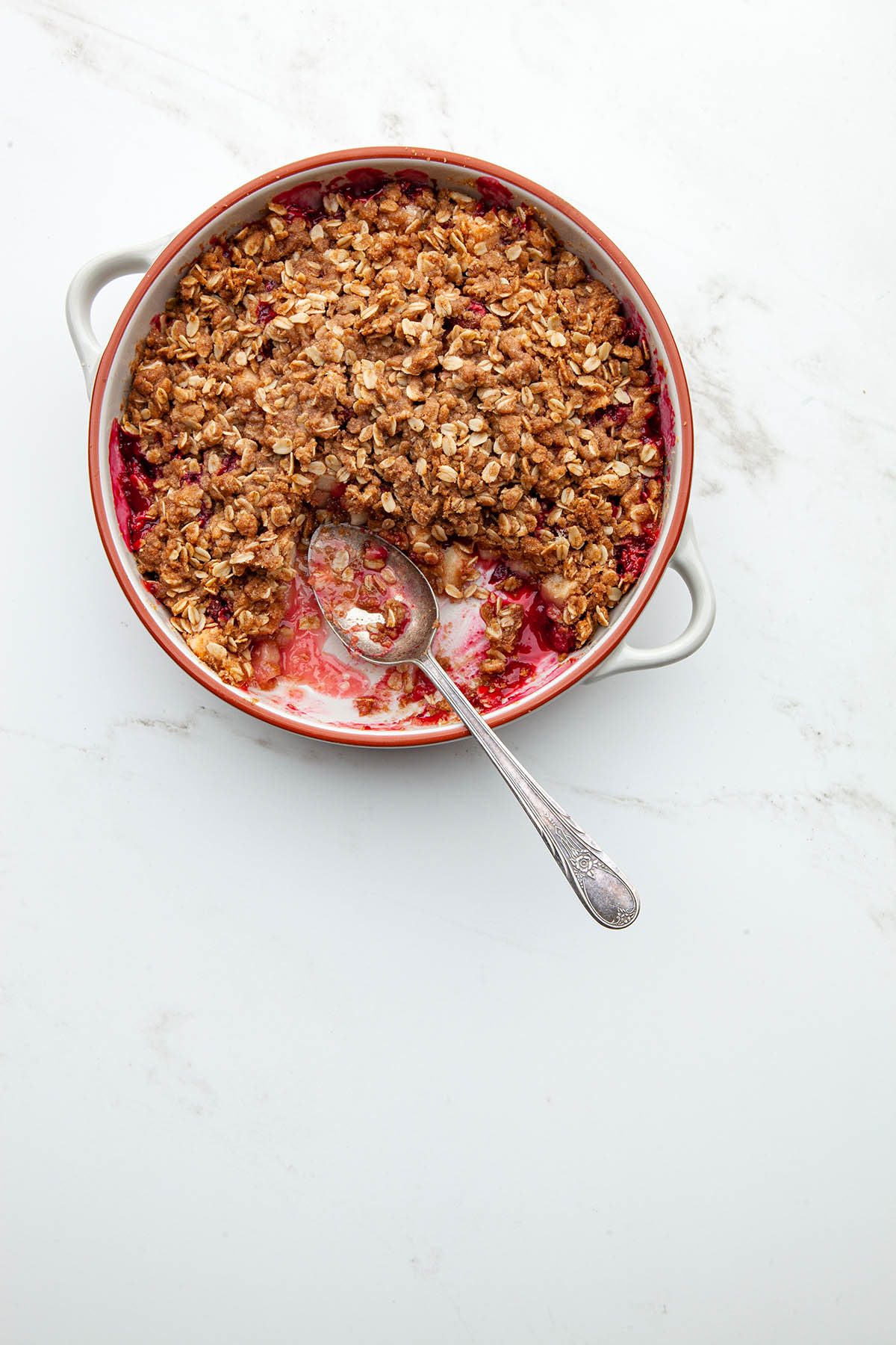 A pan of baked fruit with oat topping with one portion removed and a spoon in the pan on a marble surface.