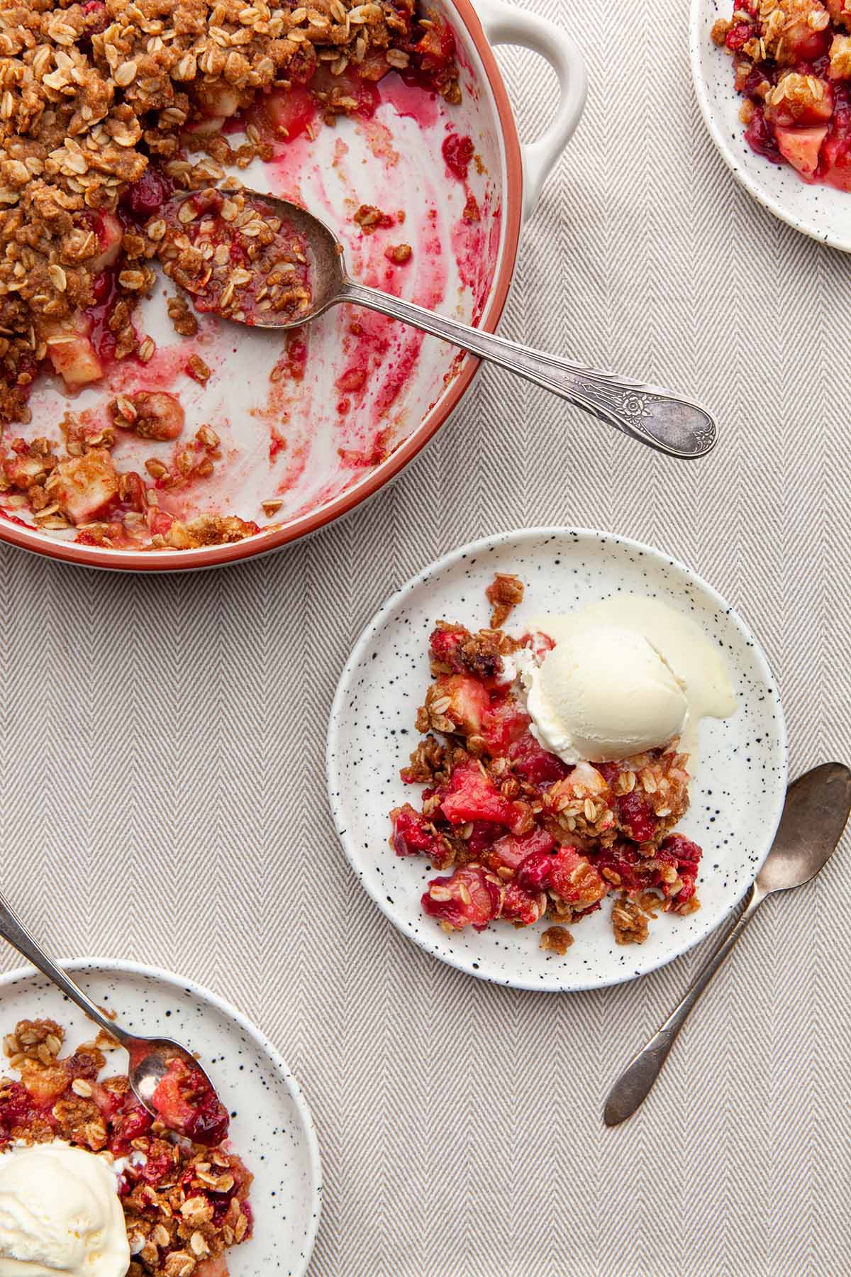 A pan of cranberry apple crisp on a table with three servings on plates topped with ice cream.