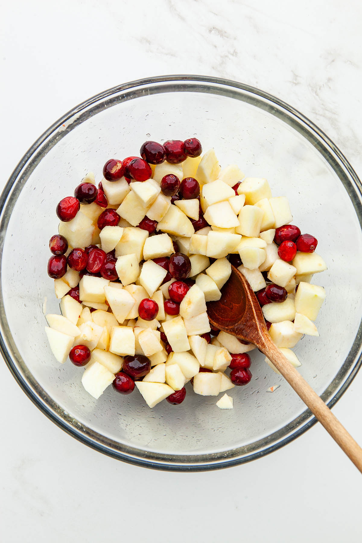 A large glass bowl of chopped apples and whole cranberries with a wooden spoon inside the bowl.