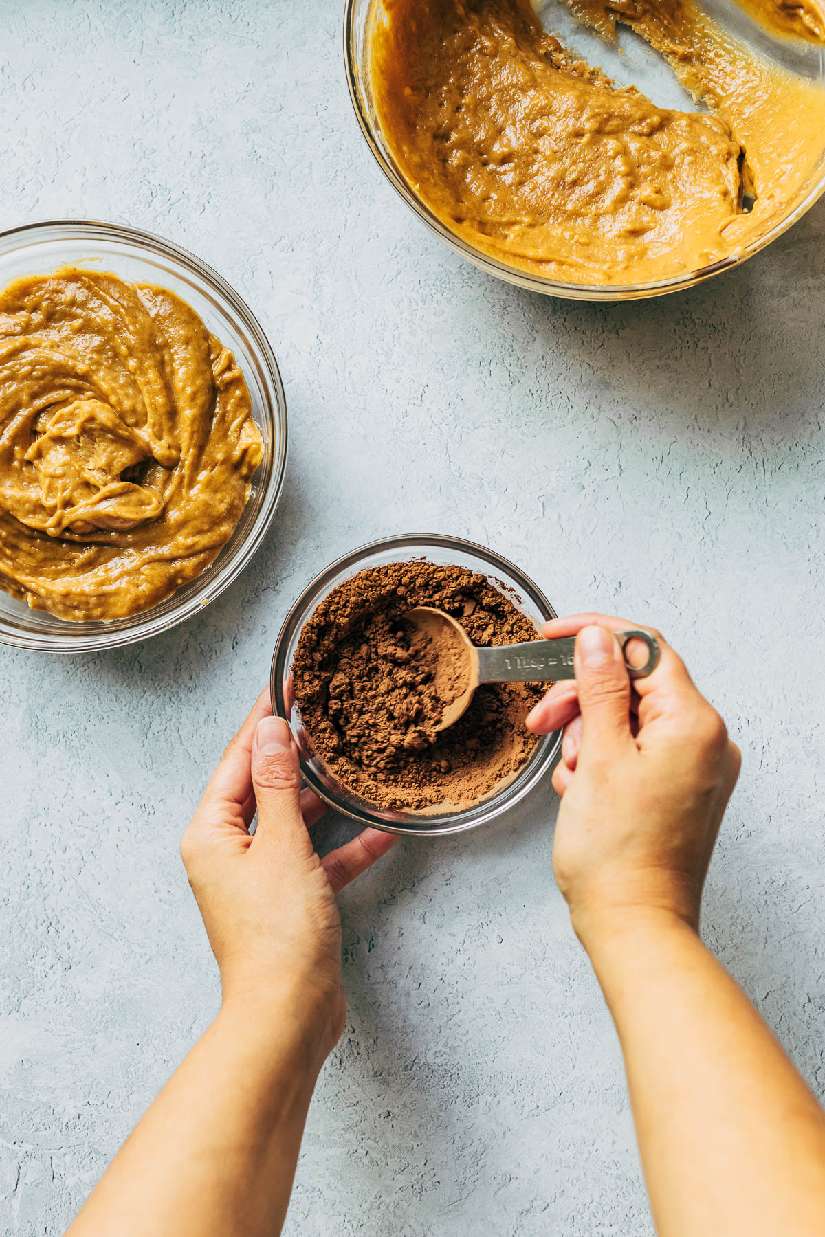 Hands scooping cocoa powder from a small bowl with a tablespoon measuring spoon.