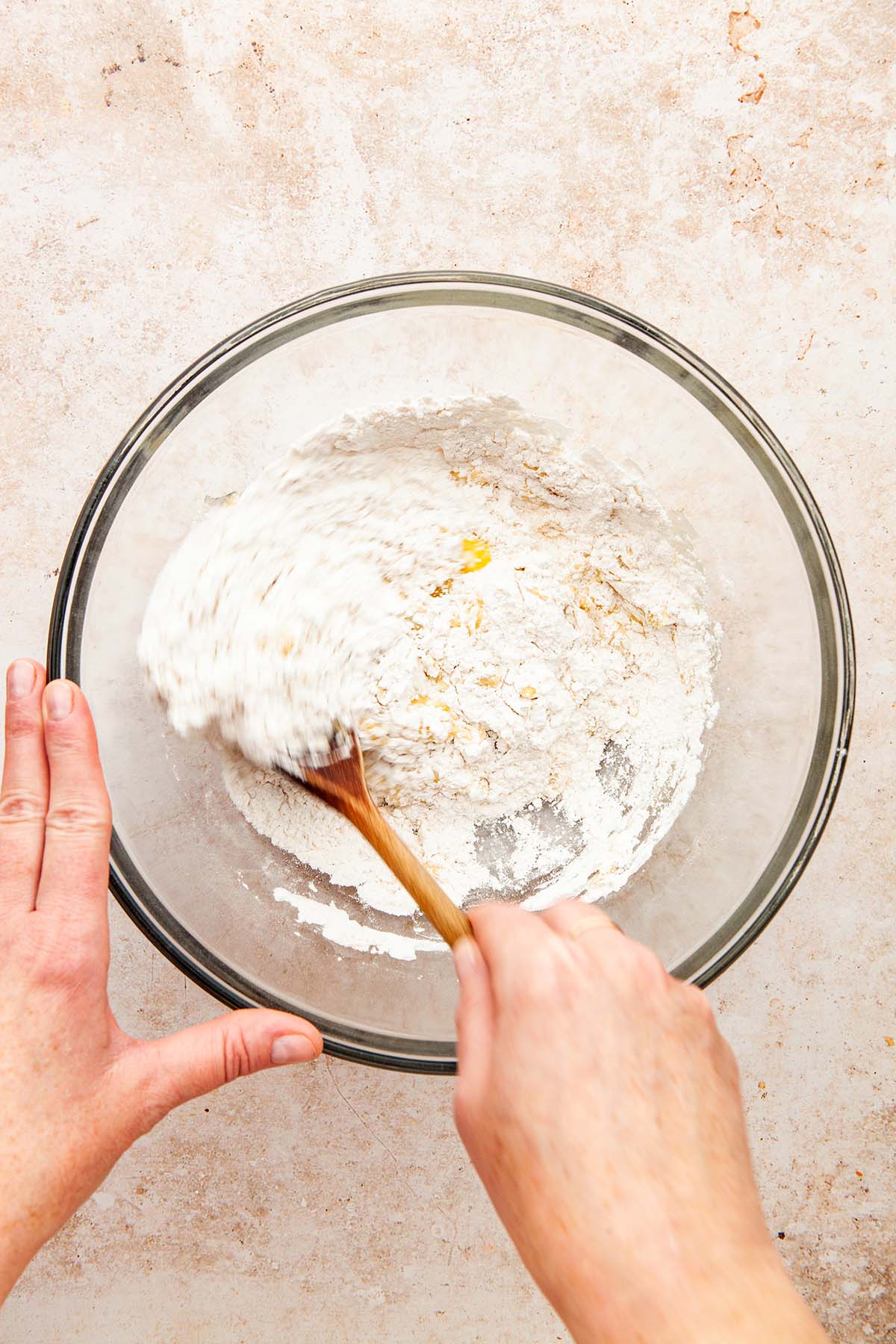 A hand mixing melted butter into flour in a glass bowl using a wooden spoon.