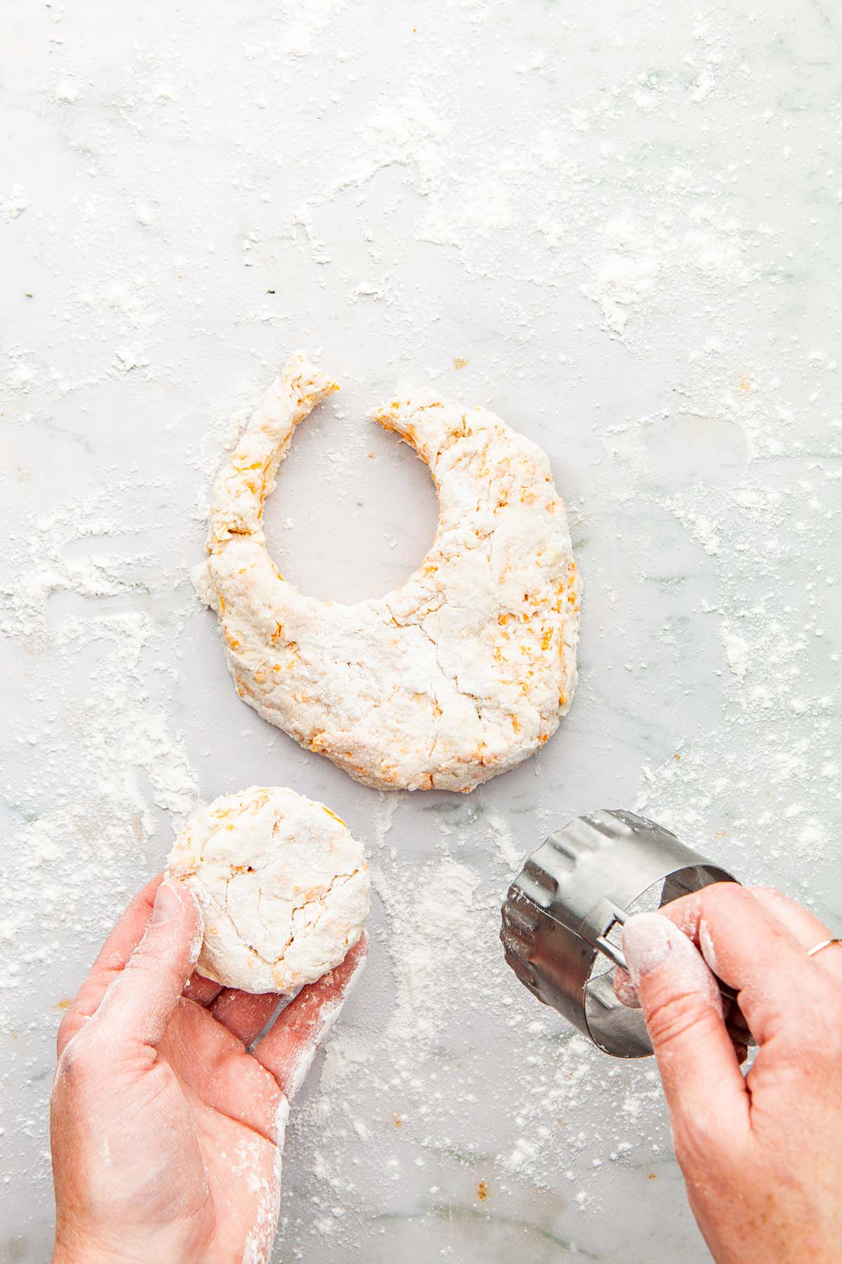 Hands holding a cut out biscuit and a biscuit cutter above rolled out dough on a marble surface.