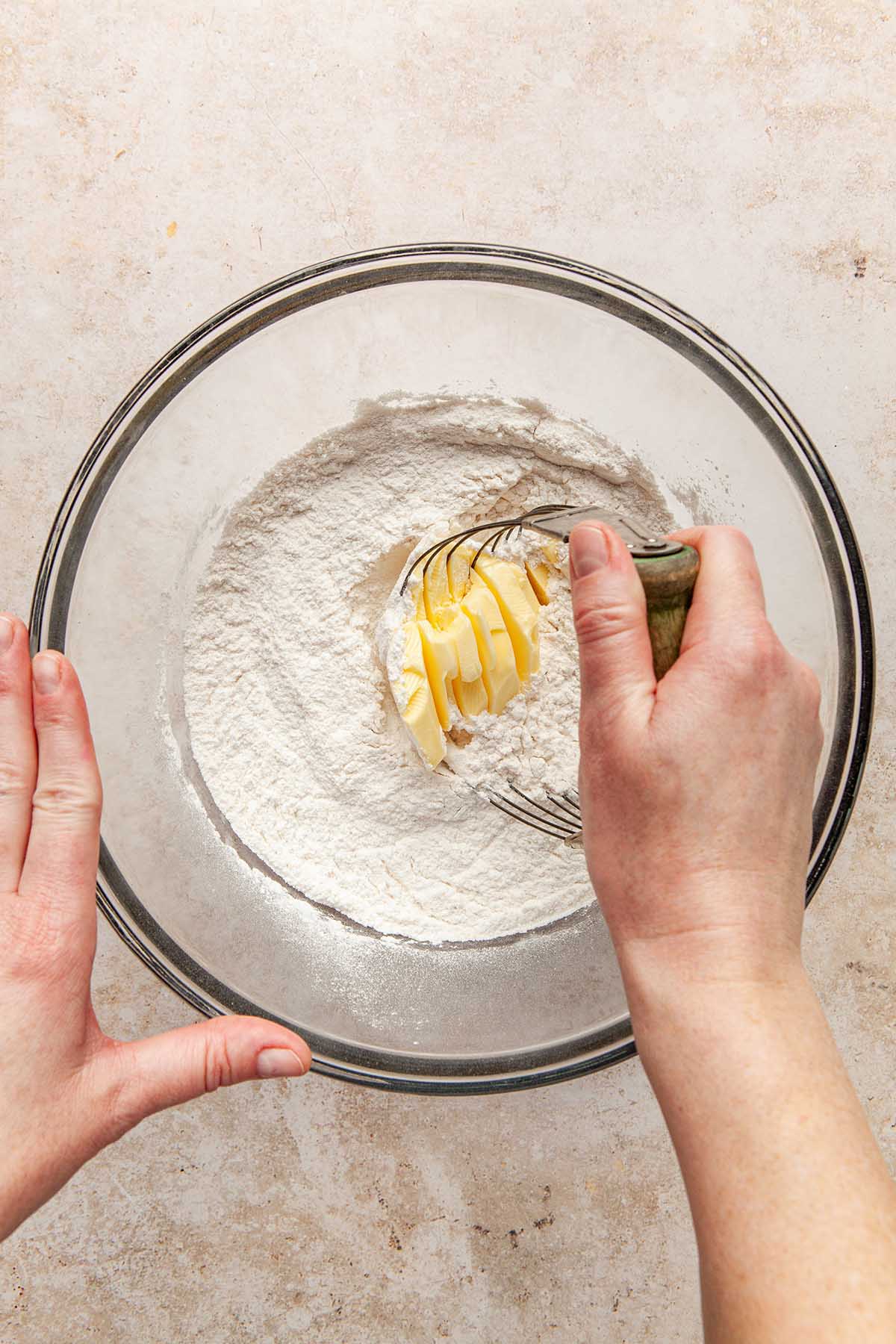 A hand holding a pastry blender, sunk into a soft chunk of butter, about to mix the butter into a bowl of flour and other dry ingredients.