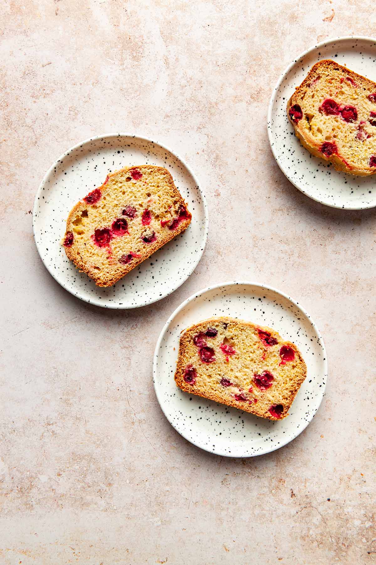 Three slices of cranberry orange loaf on small white speckled plates.