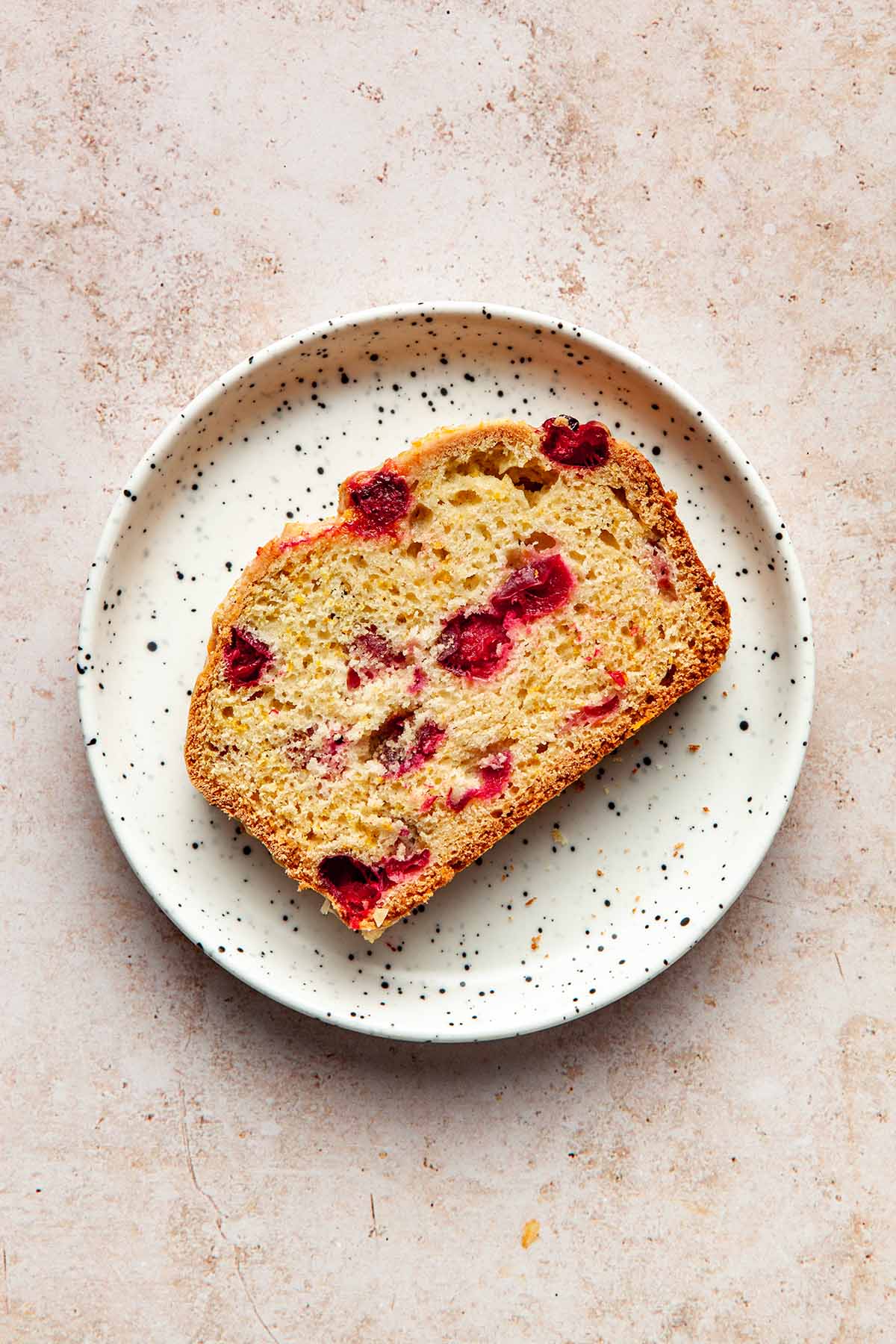 A slice of cranberry orange loaf on a small white speckled plate.