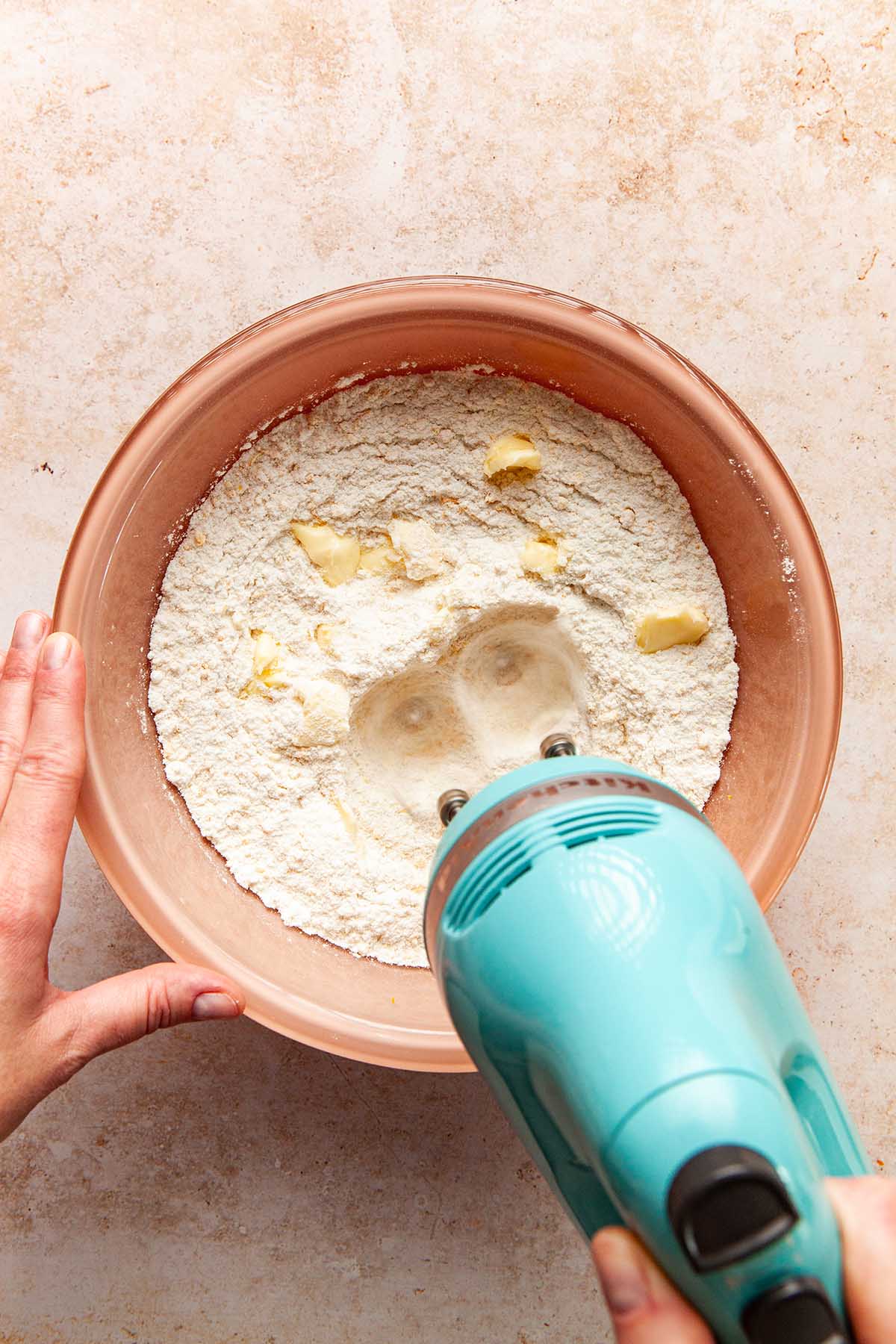 A hand using a hand mixer to mix small pieces of butter into flour.
