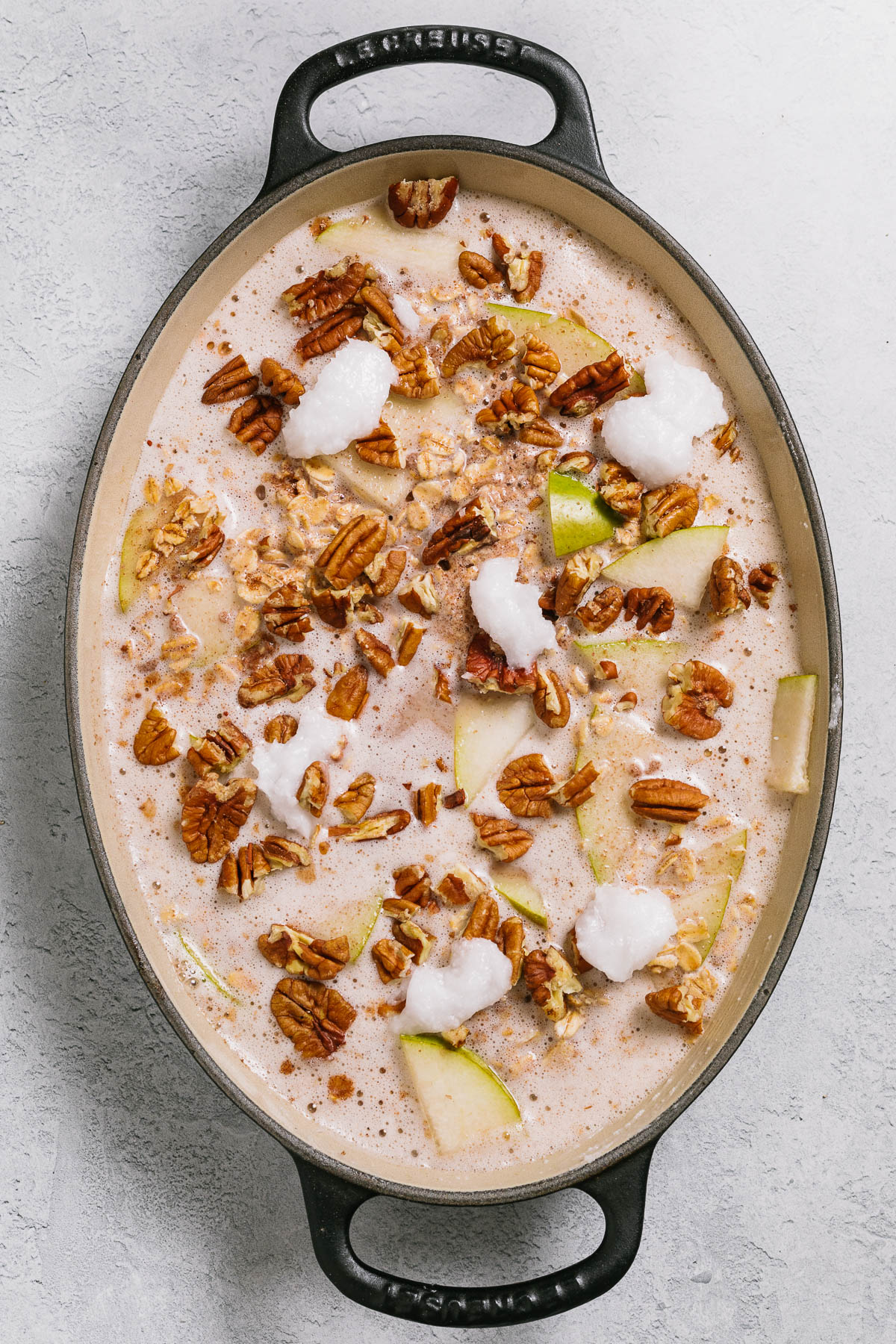 A baking dish of oats, spices, maple syrup, and almond milk with pears, chopped pecans, and chunks of solid coconut oil.