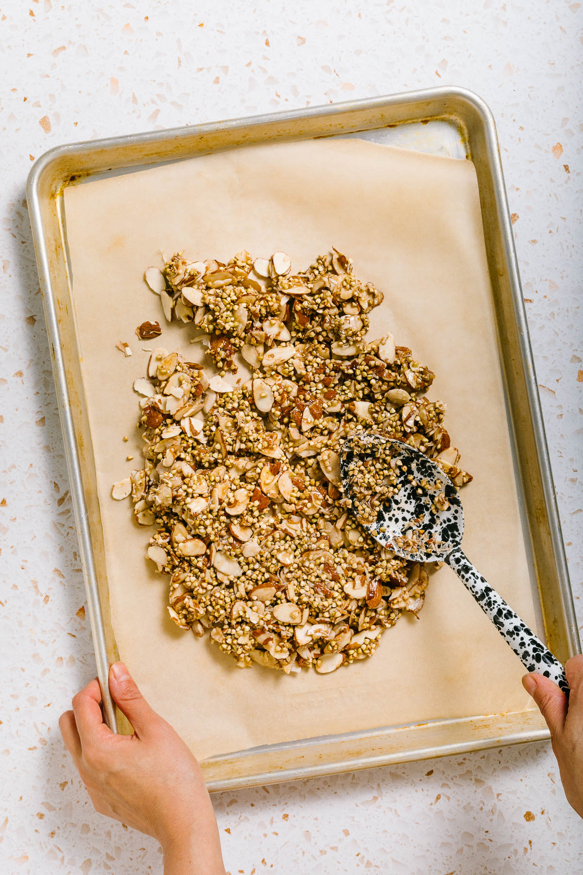 A hand using a large spoon to spread a mixture of brown rice syrup and nuts onto a baking sheet lined with parchment.