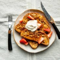 Souroudhg French toast on a plate with strawberries, maple syrup, whipped cream, and a knife and fork.