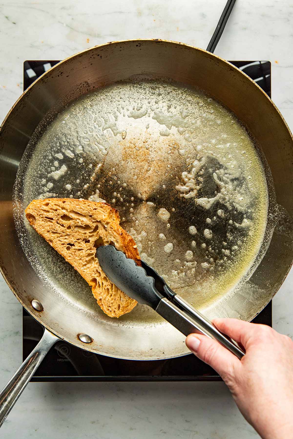 A hand using tongs to flip over a slice of bread in a frying pan.
