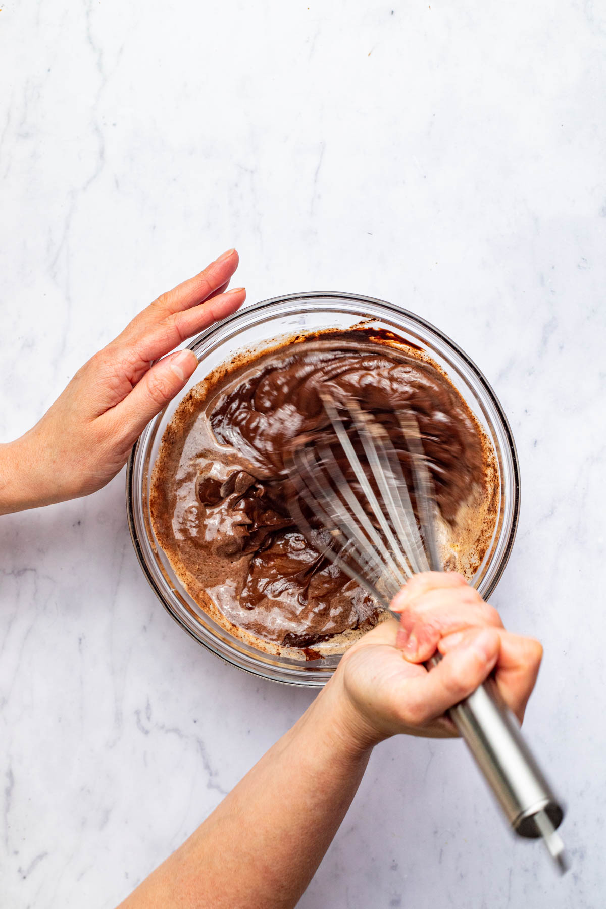 A hand using a large whisk to mix melted chocolate, butter, and cream in a glass bowl.