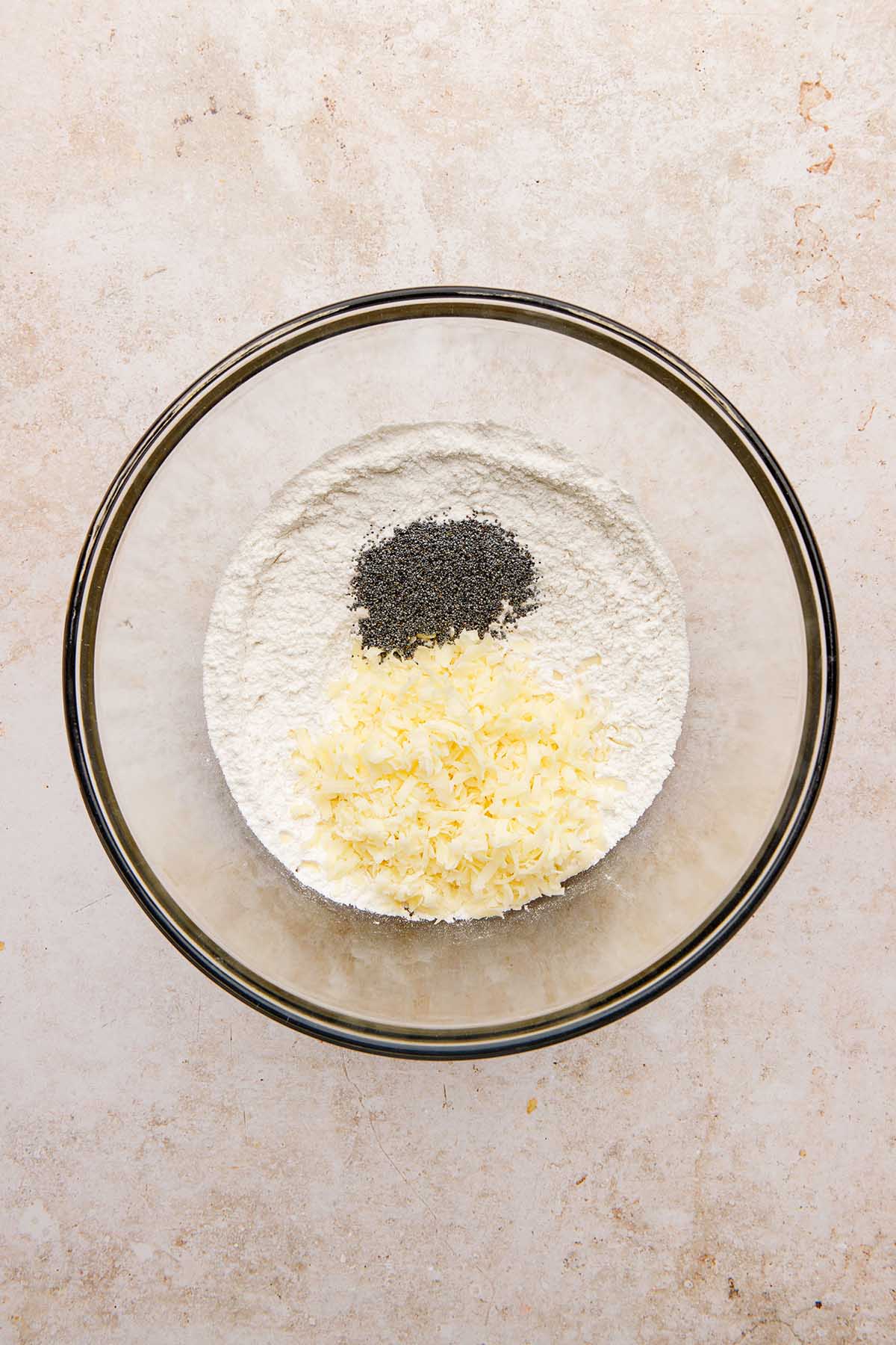 A bowl of flour with shredded cheese and poppy seeds on top.