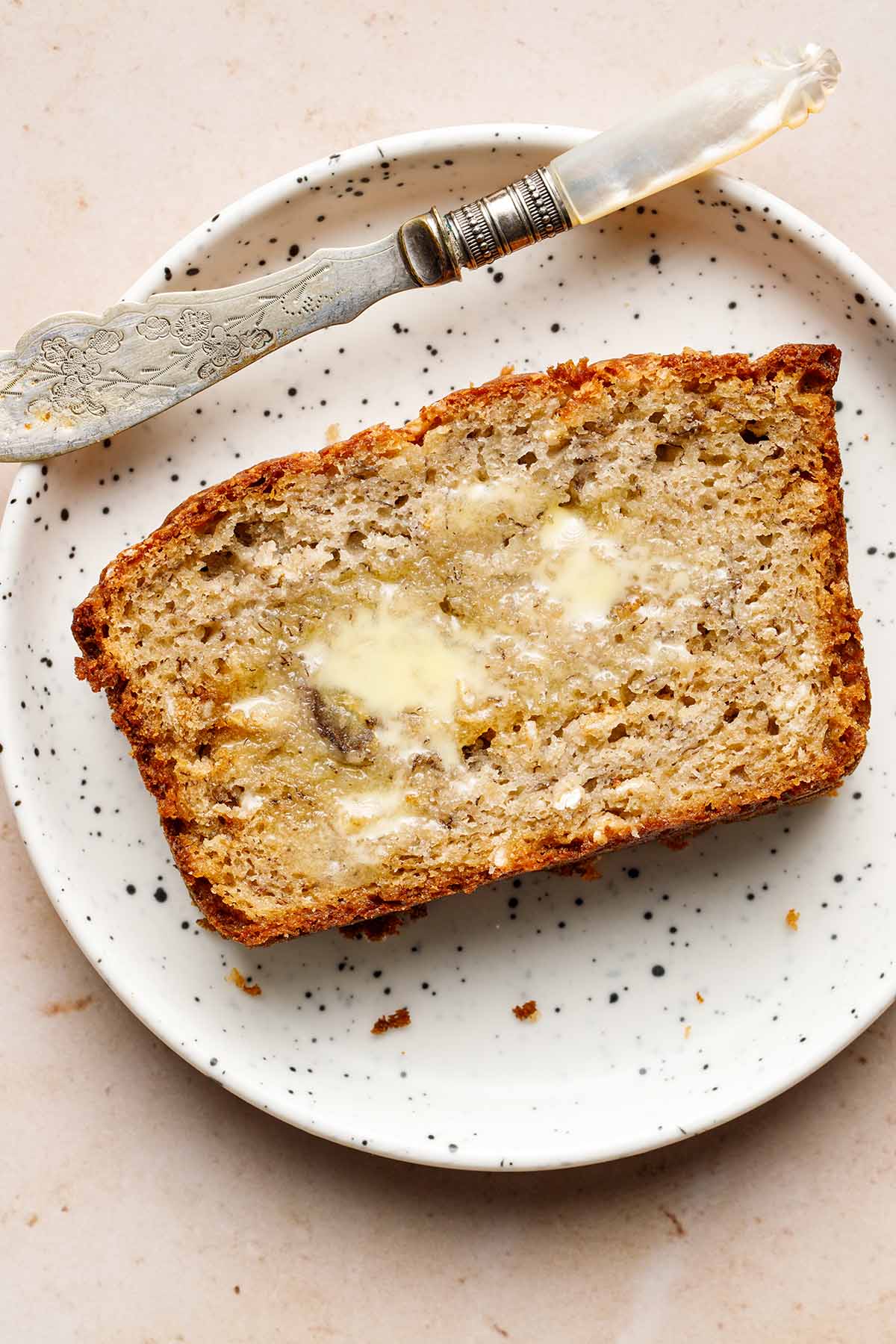 A slice of buttered sourdough banana bread on a small white speckled plate.