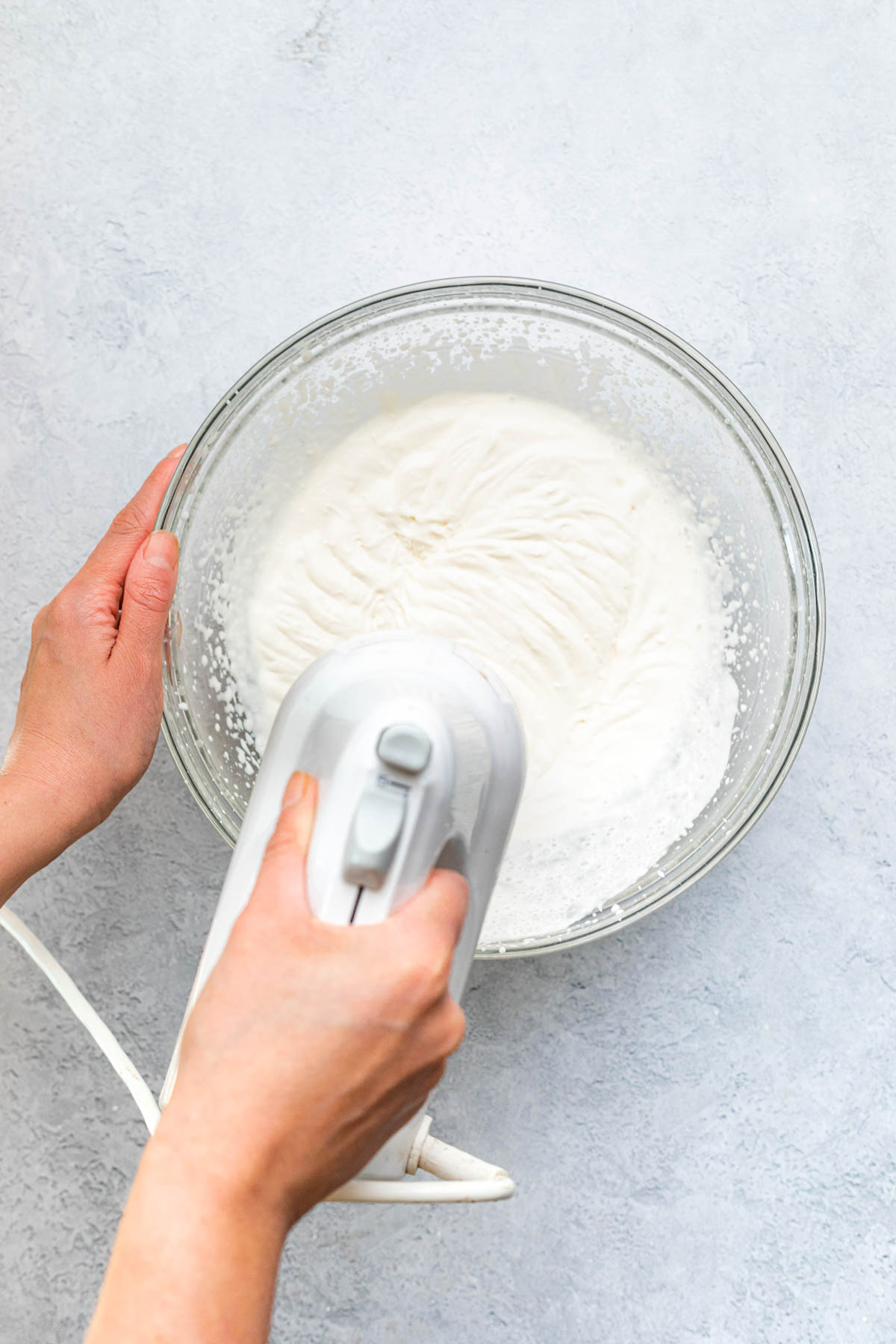 A hand using a hand mixer to whip cream in a large glass bowl.