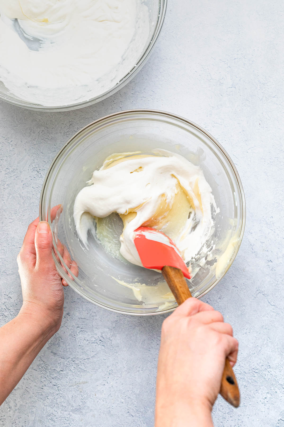 A hand folding whipped cream and melted white chocolate together in a large glass bowl.
