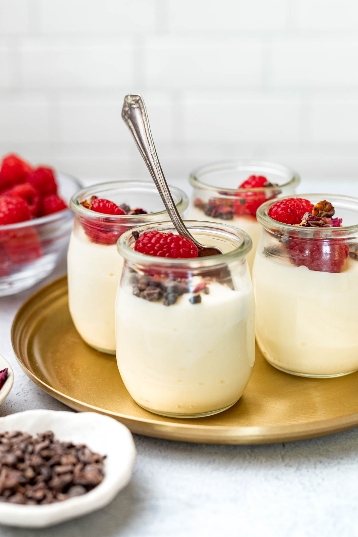 Jars of white chocolate mousse garnished with cacao nibs, raspberries, and dried rose petals on top.