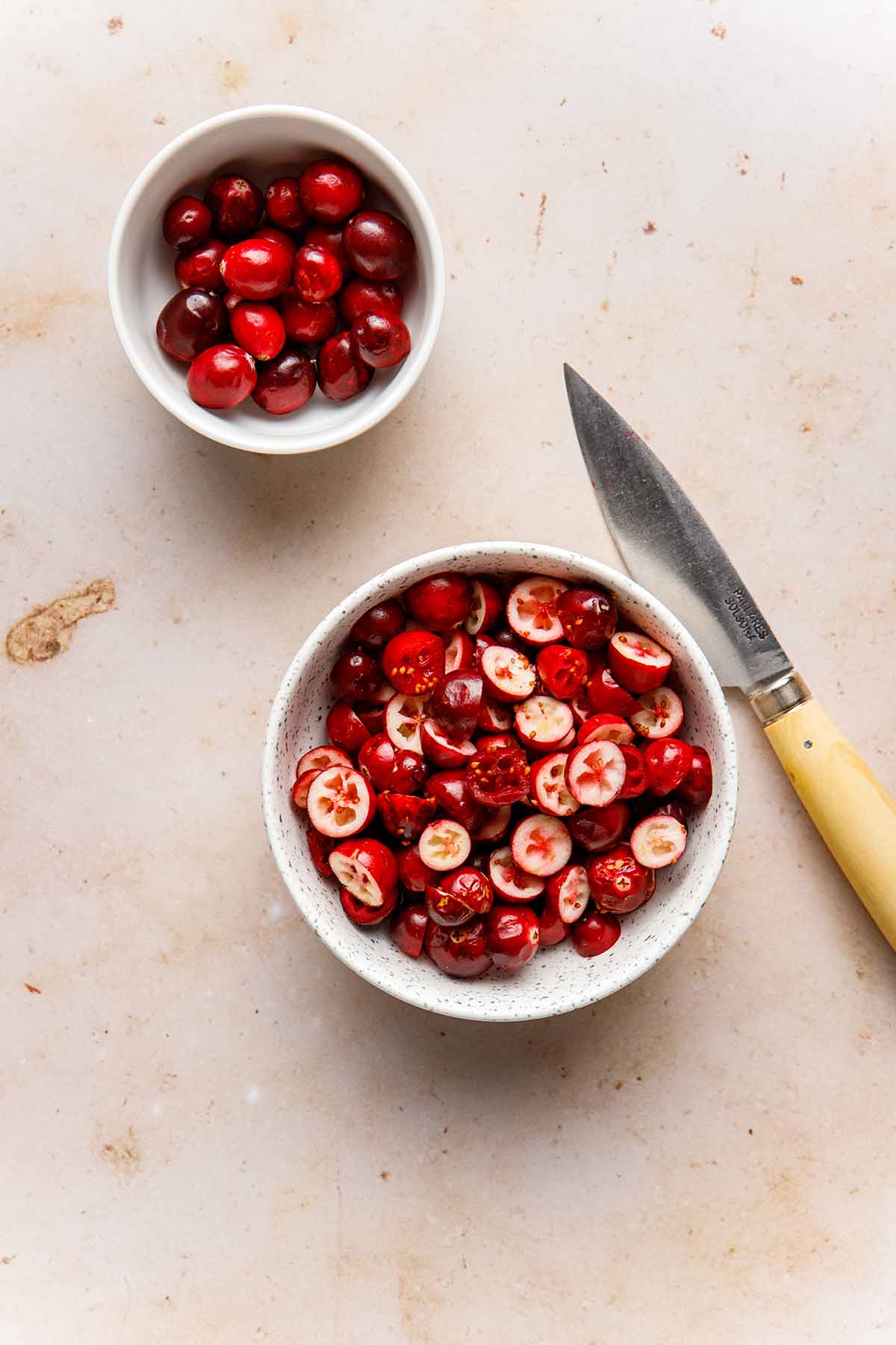 A small bowl of halved cranberries next to a knife and a smaller bowl of whole cranberries.