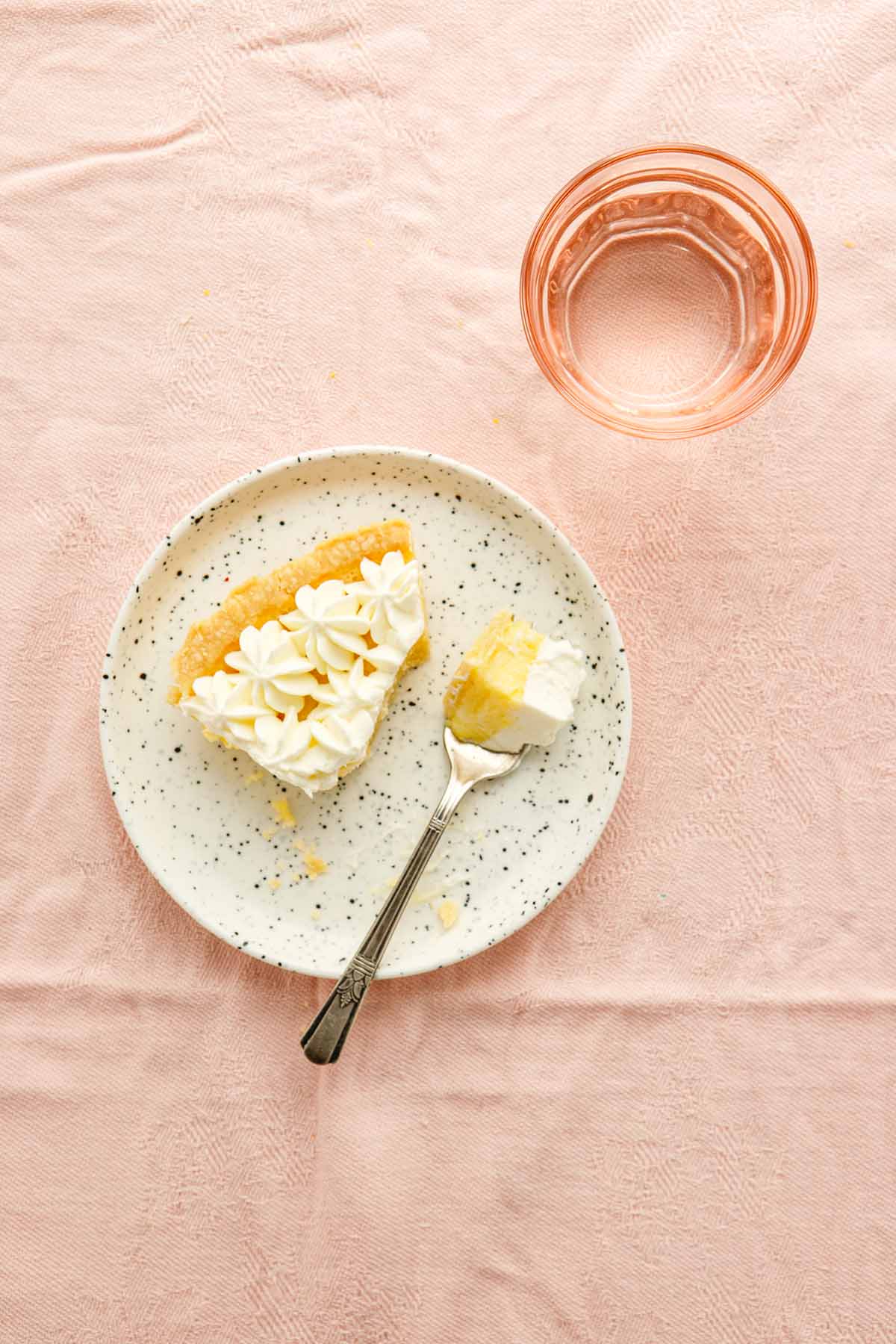 A slice of lemon chess pie on a small white plate on a table covered in pink linen with a pink glass filled with water nearby.