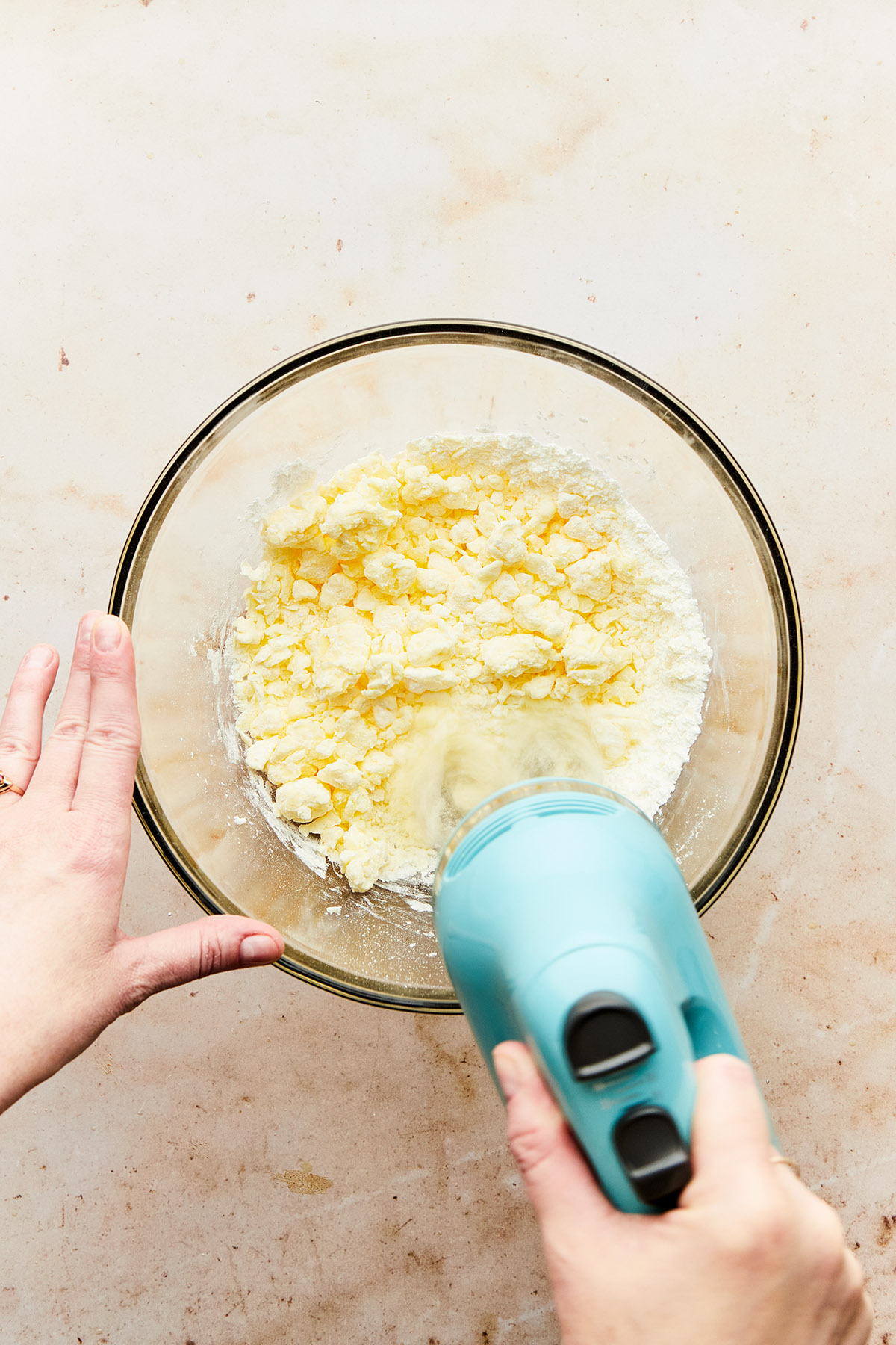 A hand using a hand mixer to whip butter and powdered sugar together in a large glass bowl.