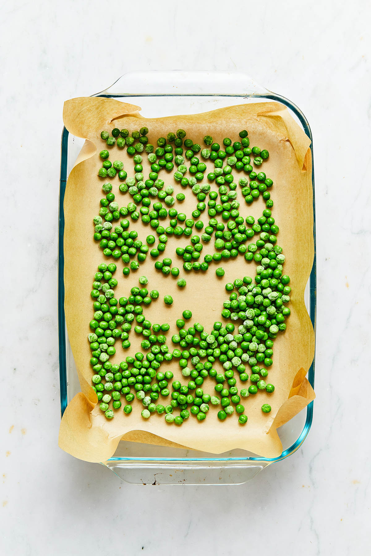 A baking dish lined with parchment paper with green peas scattered over the bottom of the dish.