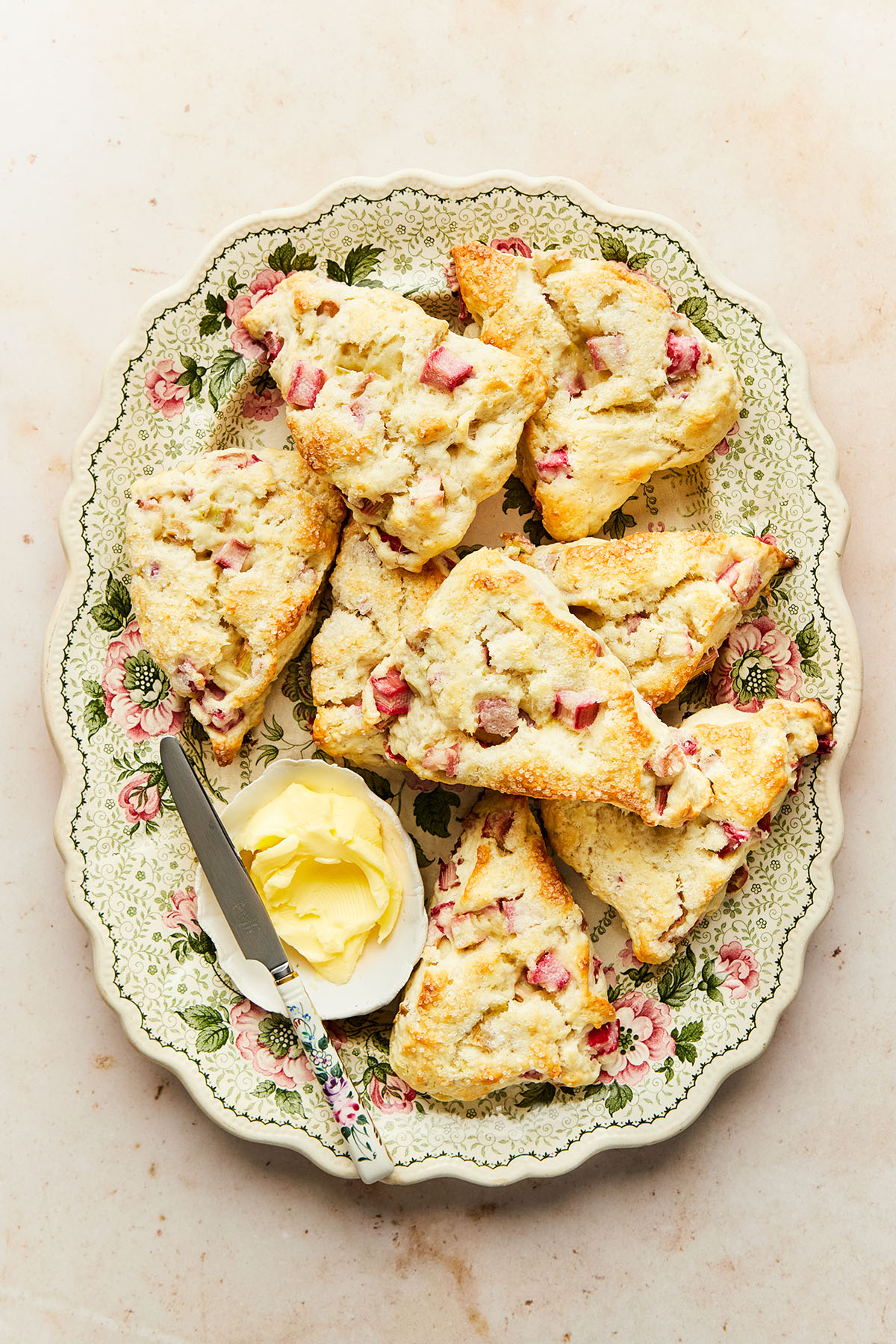 A pink and green flowered platter of rhubarb scones with a small dish of butter and a knife.