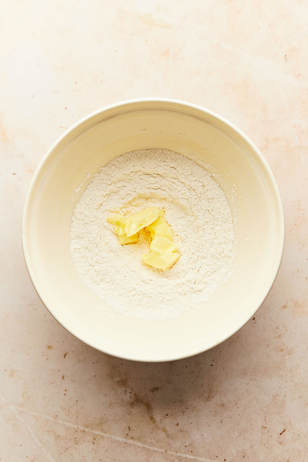 Chunks of butter on top of mixed dry ingredients in a large cream-coloured bowl.