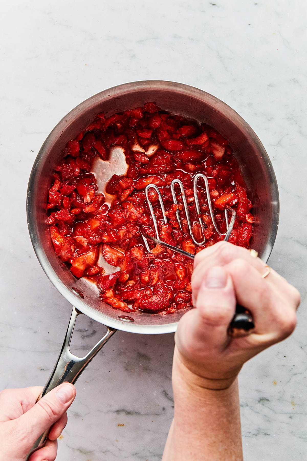 A hand using a potato masher to crush strawberries in a pot.